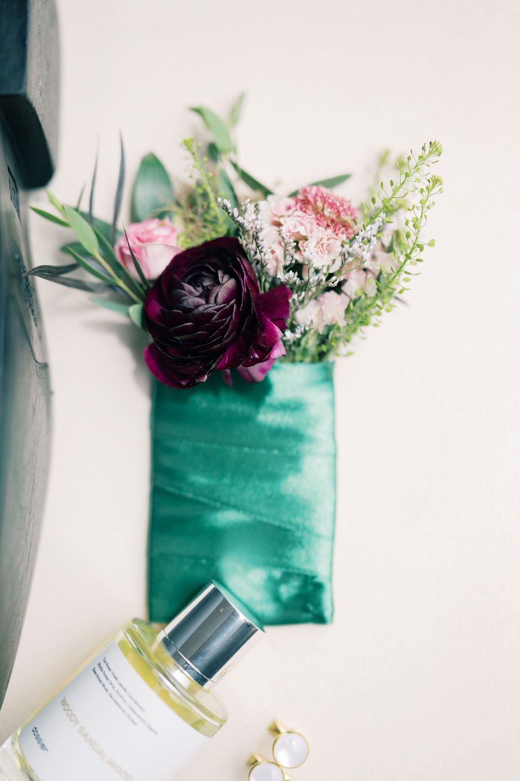 Elegant flat lay with burgundy and pink floral bouquet on green velvet ribbon, perfume bottle, and gold ring on white background.
