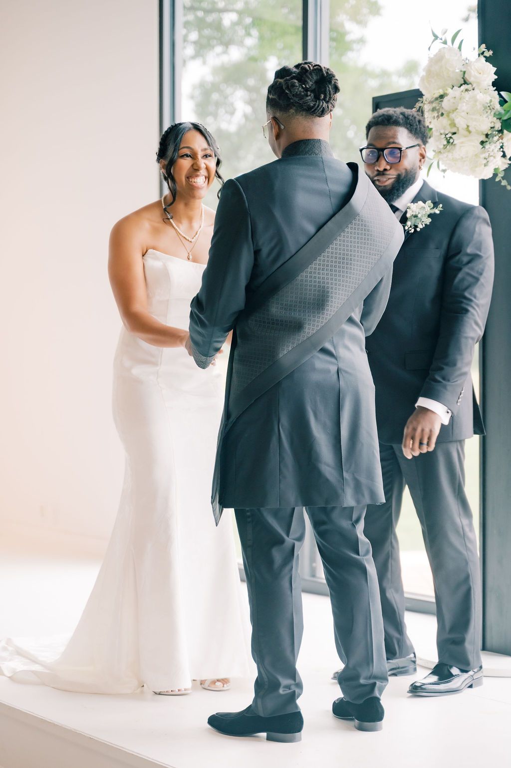 Bride in white gown exchanges vows with groom in gray suit, officiant presiding at elegant indoor wedding ceremony.