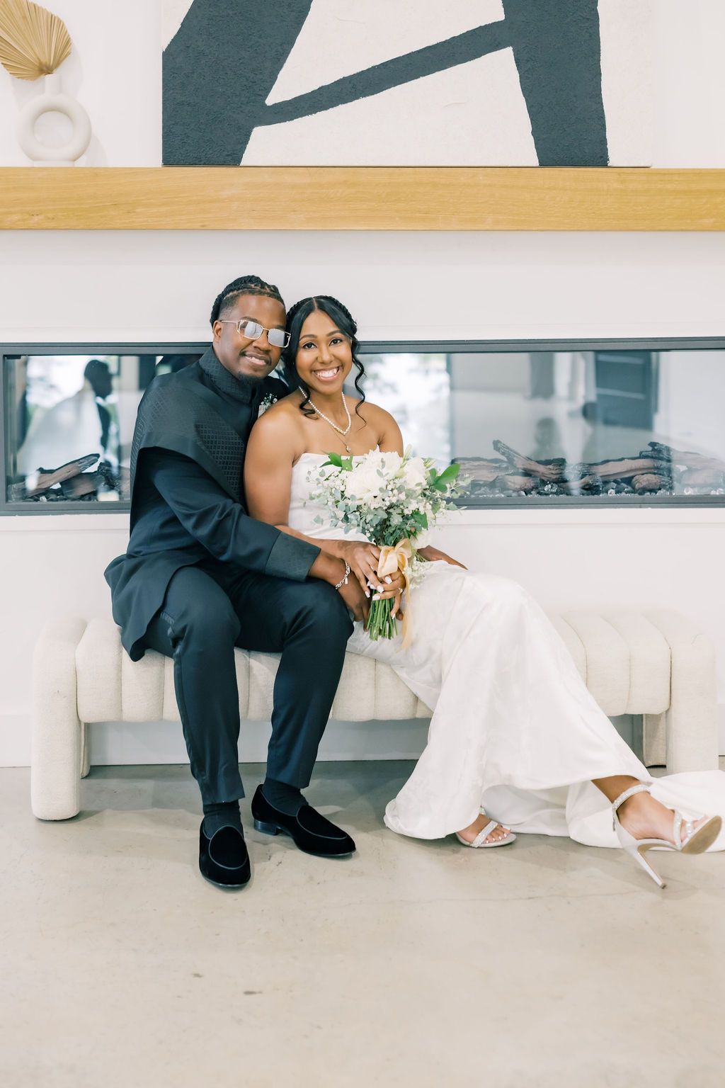 A smiling bride and groom sit together on a white bench, she holds a bouquet in a wedding gown, he wears a dark suit.