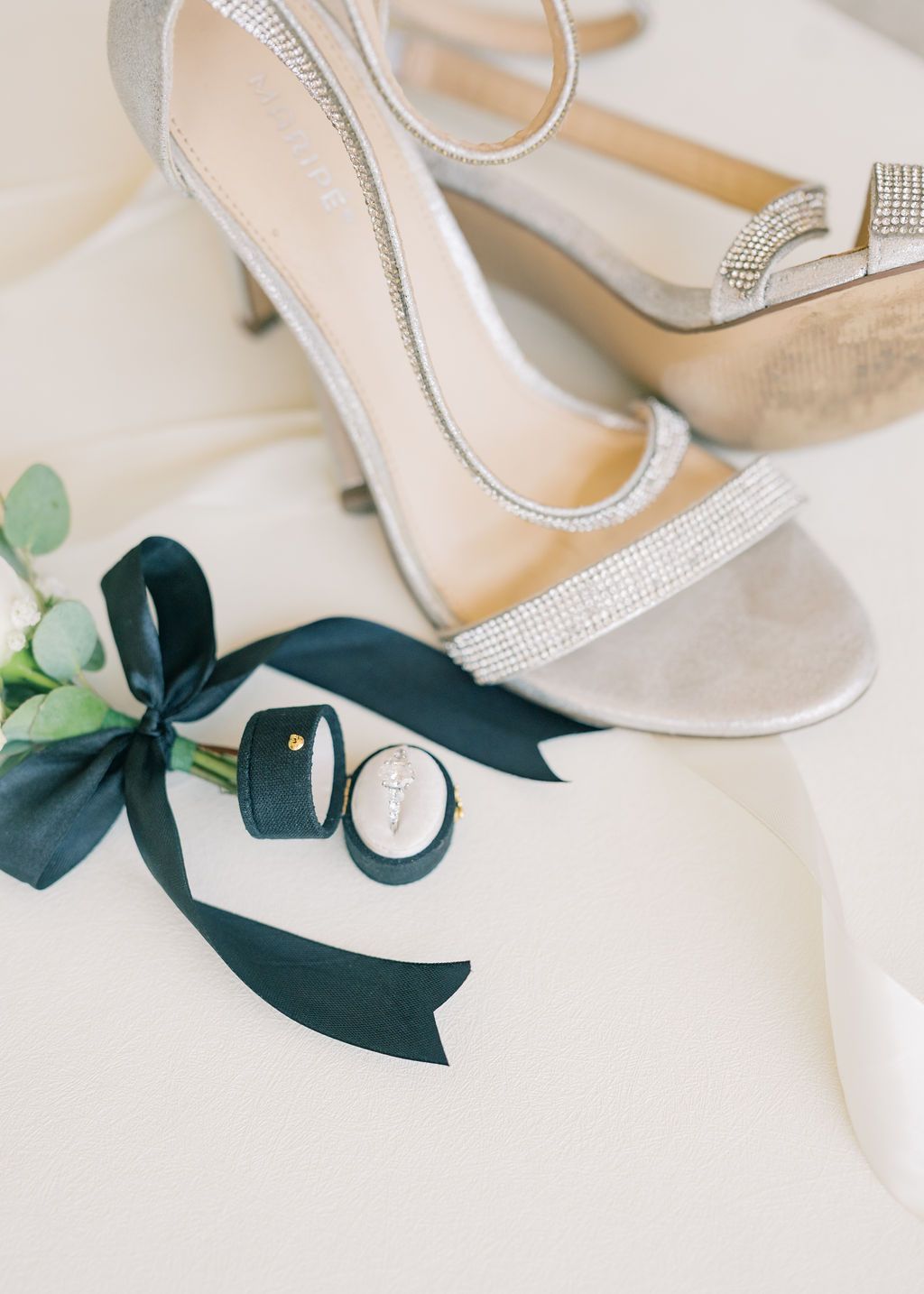 Silver rhinestone ankle strap heels beside a dark green velvet ring box with ribbon and boutonniere on white fabric.