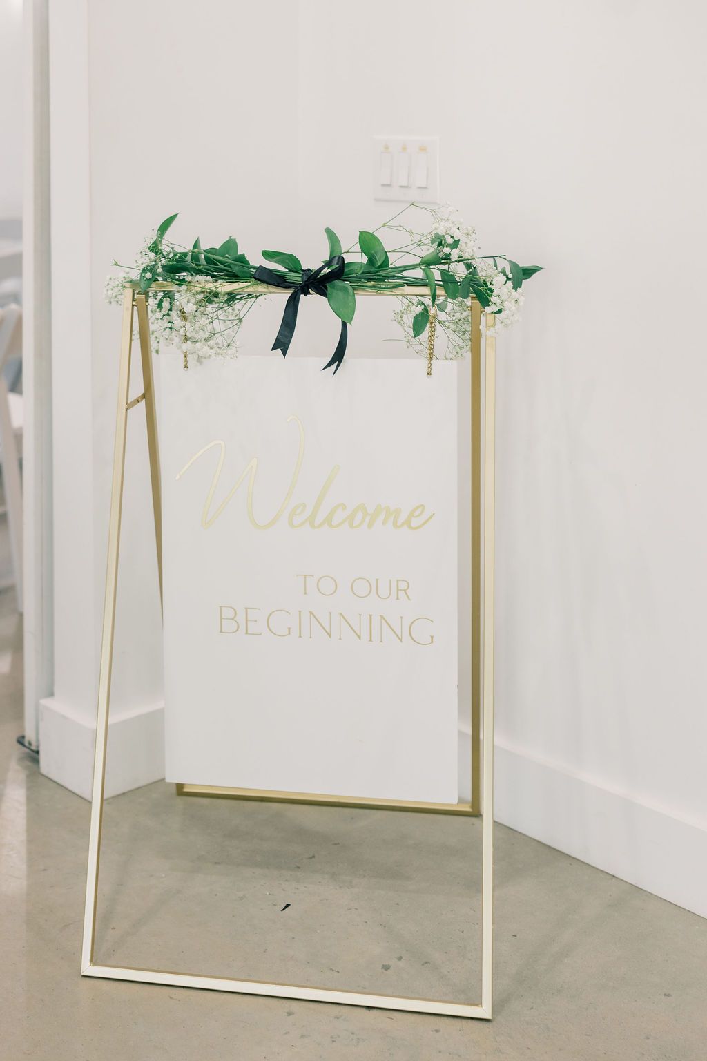 Gold-framed wedding welcome sign reading Welcome To Our Beginning decorated with greenery and ribbon.