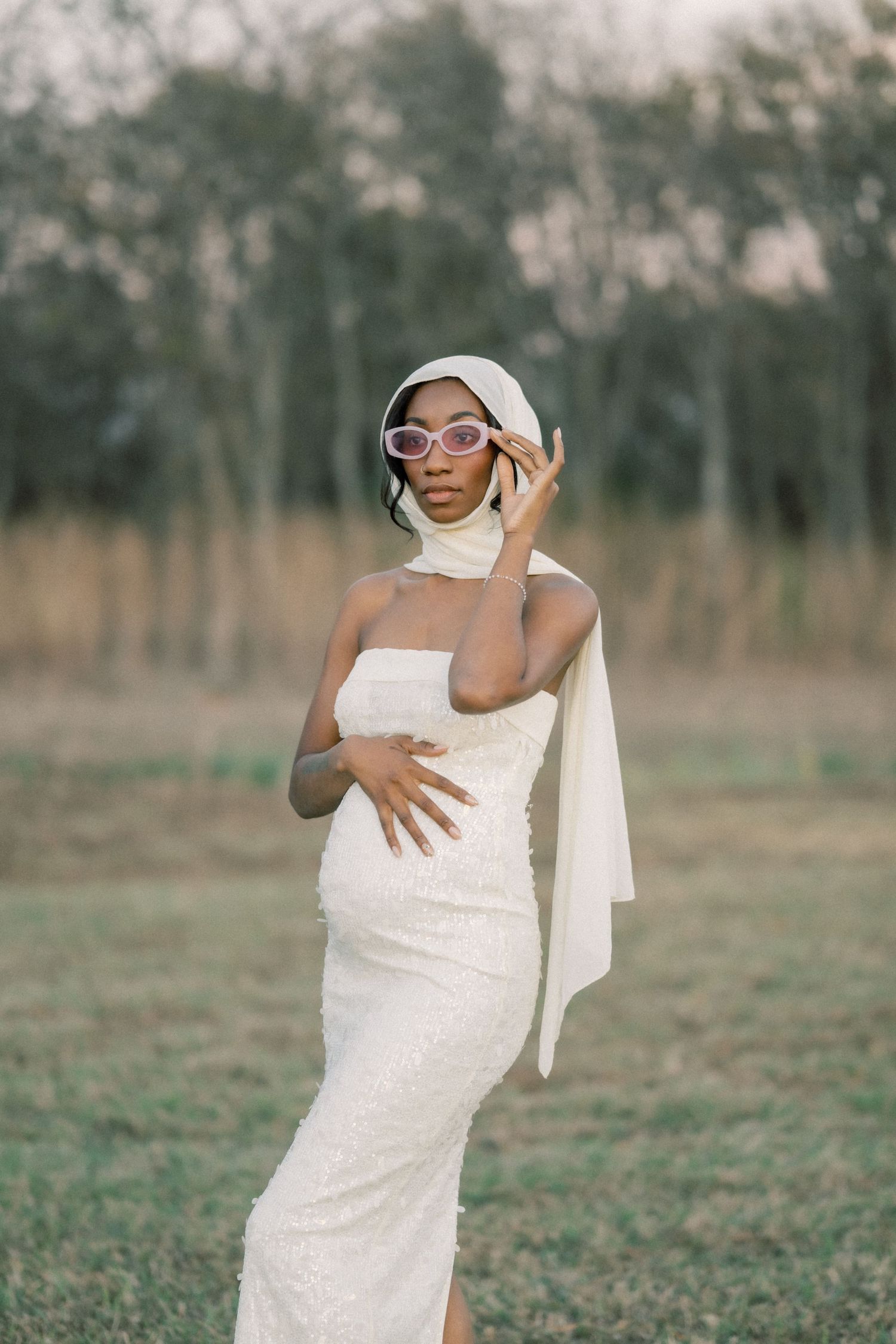 Person in a white dress poses playfully in a field with sunglasses and a hat.
