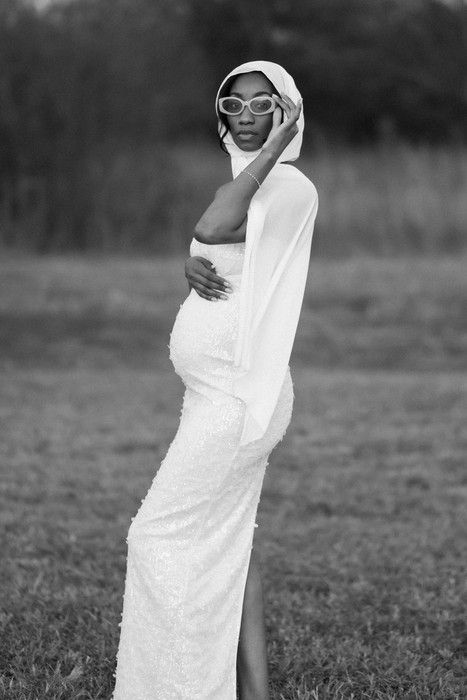 Black and white maternity photo showing woman in white dress and sunglasses in field.