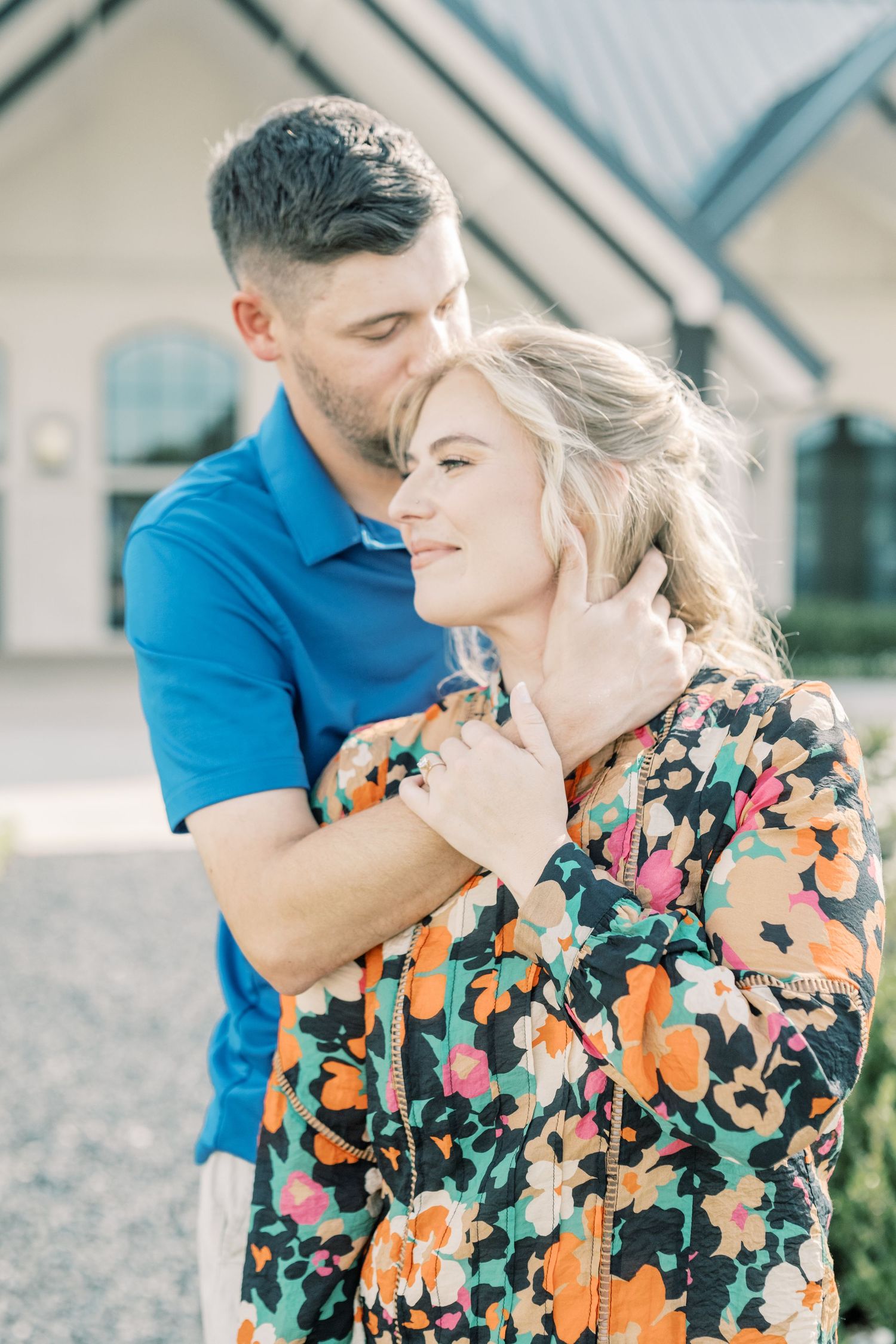 A man in a blue shirt tenderly kisses a smiling woman in a vibrant floral dress outdoors.