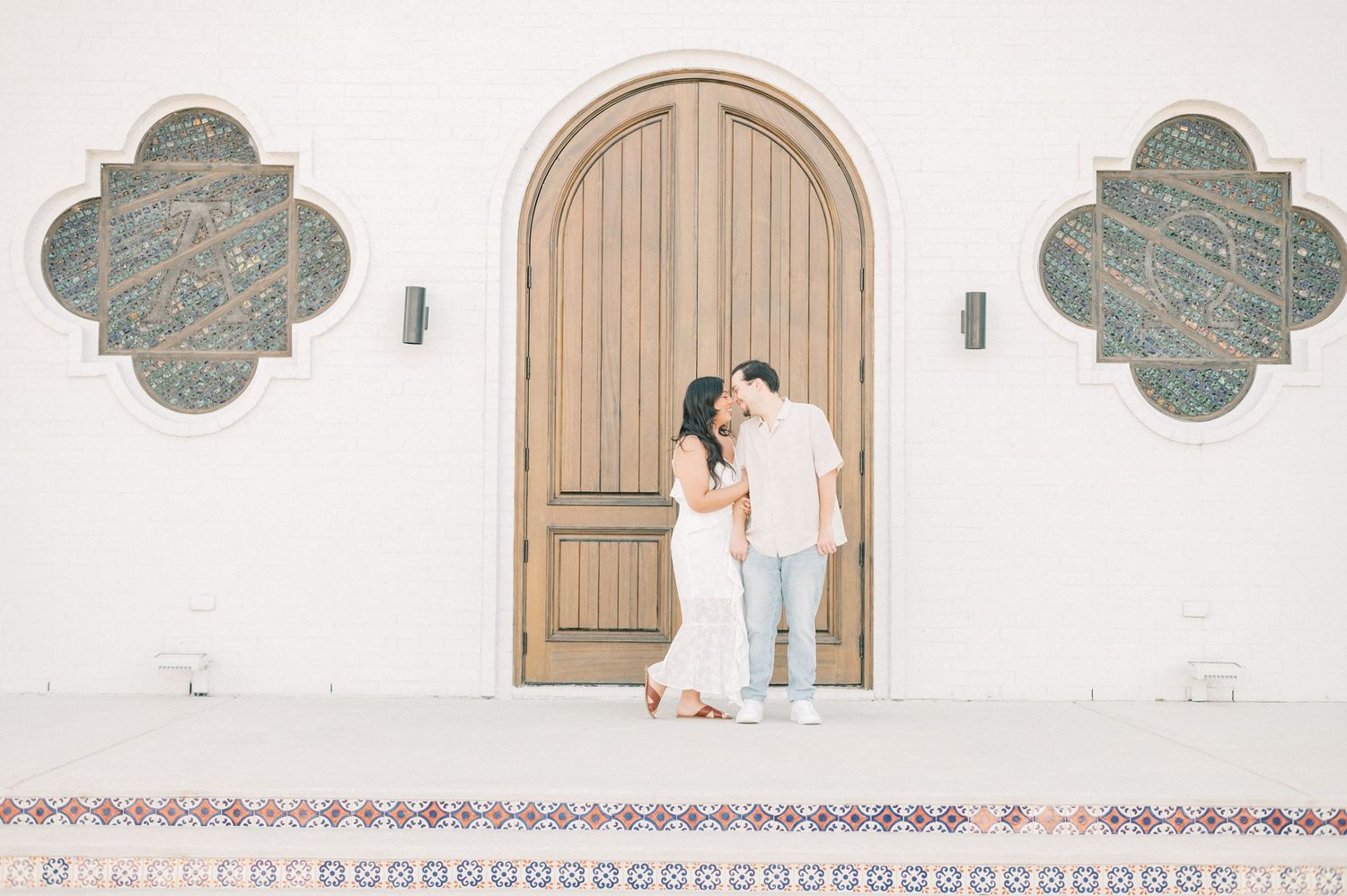 A couple embraces in front of a Spanish colonial style white wall with arched wooden doors and decorative details.