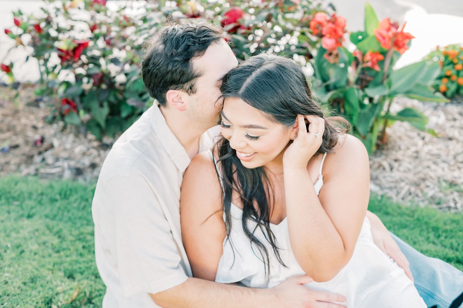 A couple shares an intimate moment in a garden setting with red flowers in the background.