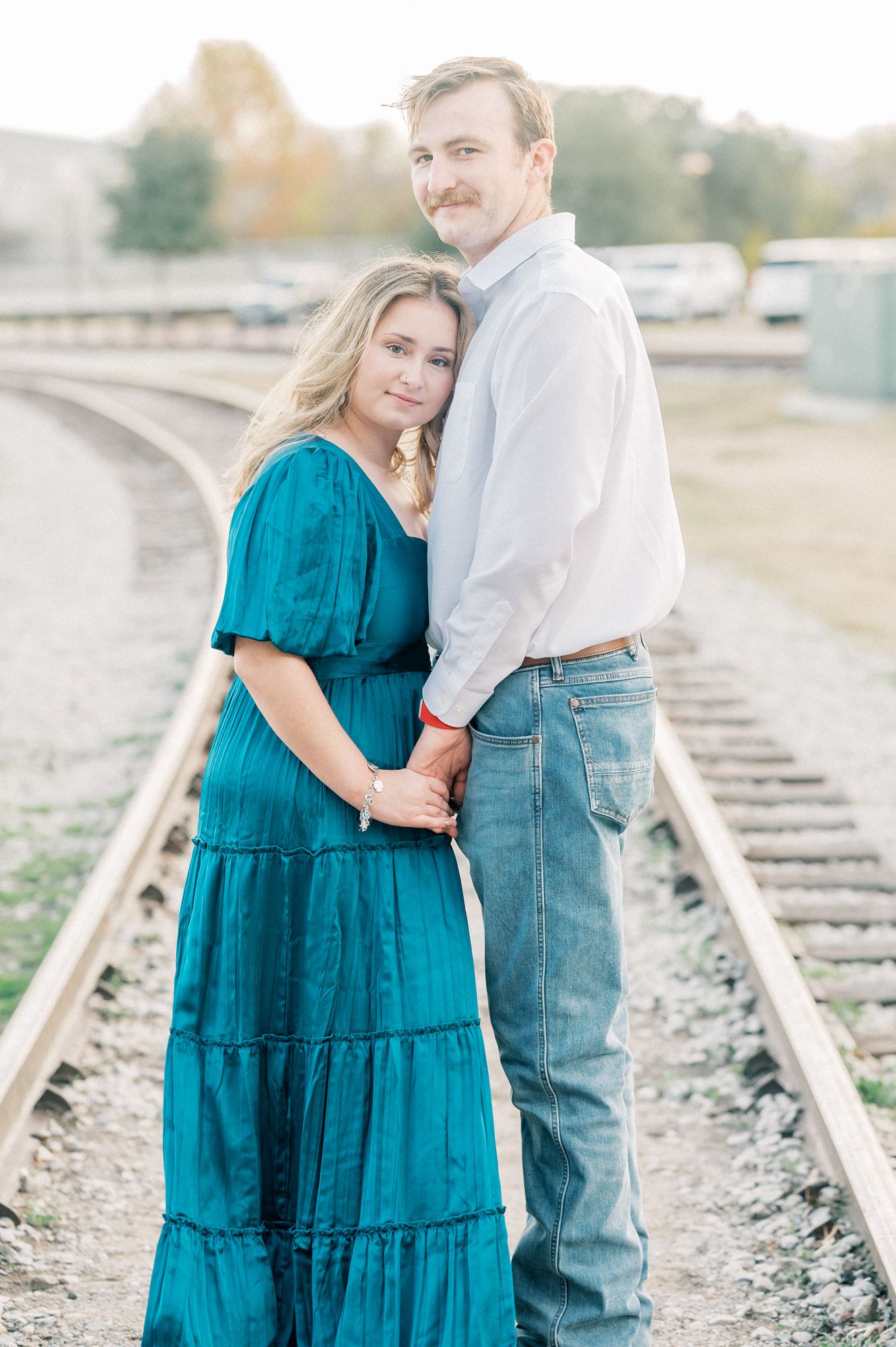 Couple poses together on train tracks with woman in teal dress and man in white shirt and jeans.