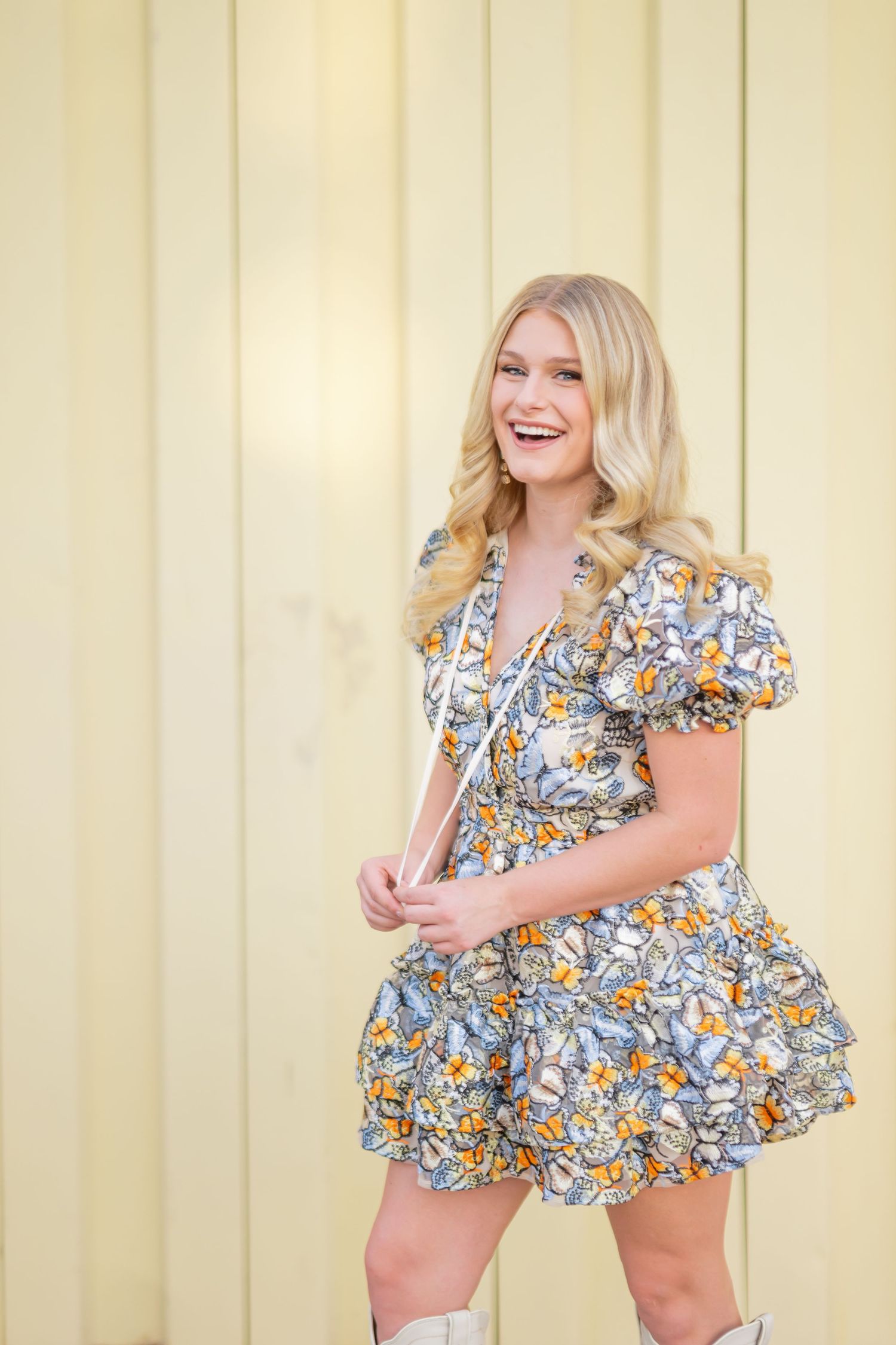 Smiling blonde woman in floral mini dress and white boots posing against a yellow striped wall.