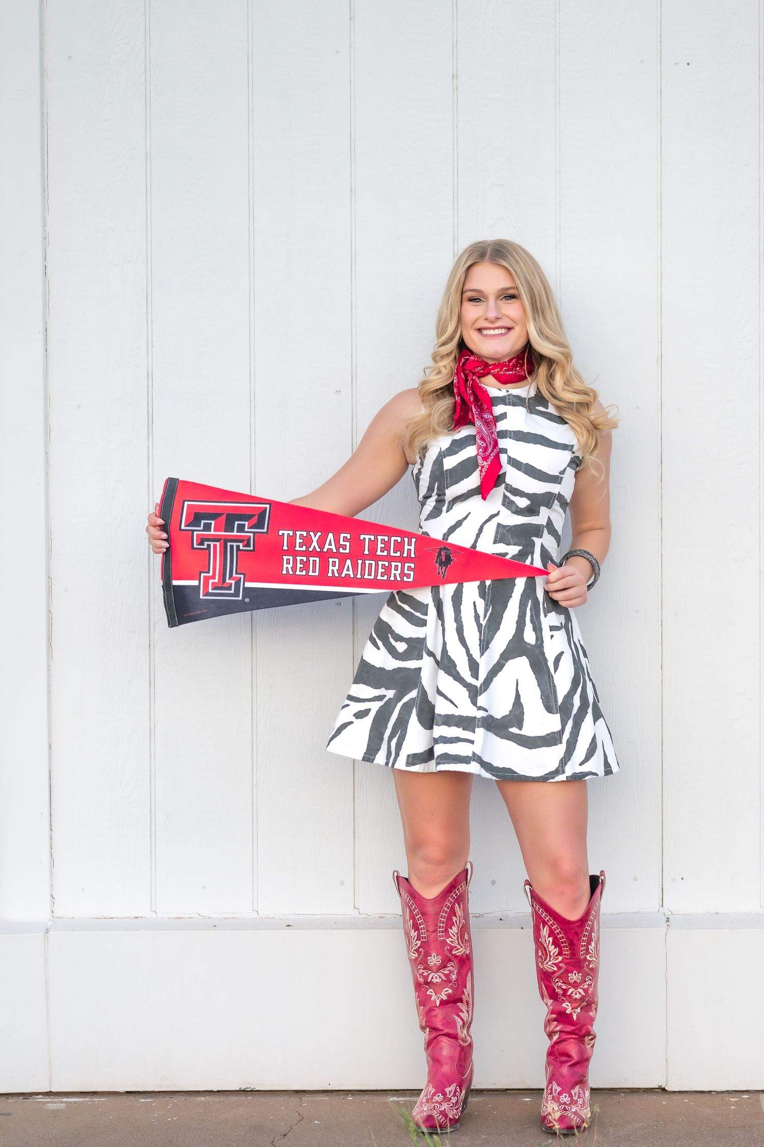Smiling blonde woman in zebra print dress and pink cowboy boots holding a Texas Tech Red Raiders pennant flag.