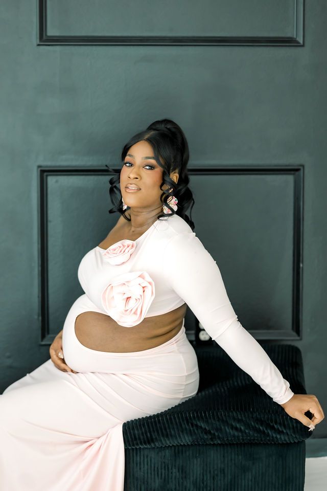 Pregnant woman in elegant white cutout maternity gown sitting gracefully against dark teal wall.