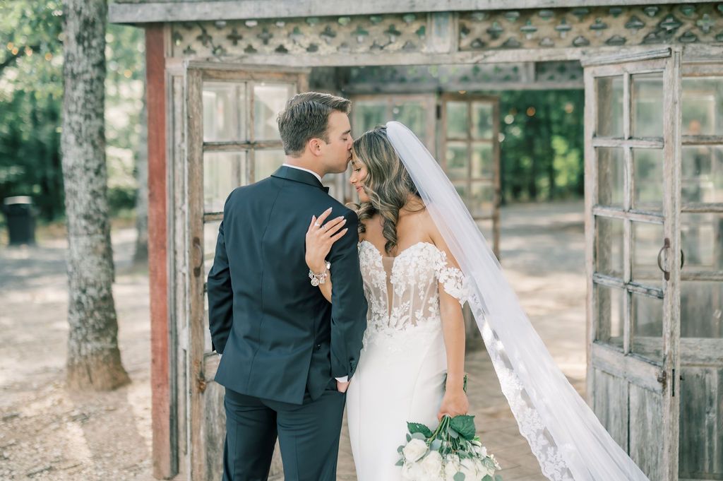 Bride and groom share a romantic kiss in front of rustic wooden doors surrounded by lush greenery.