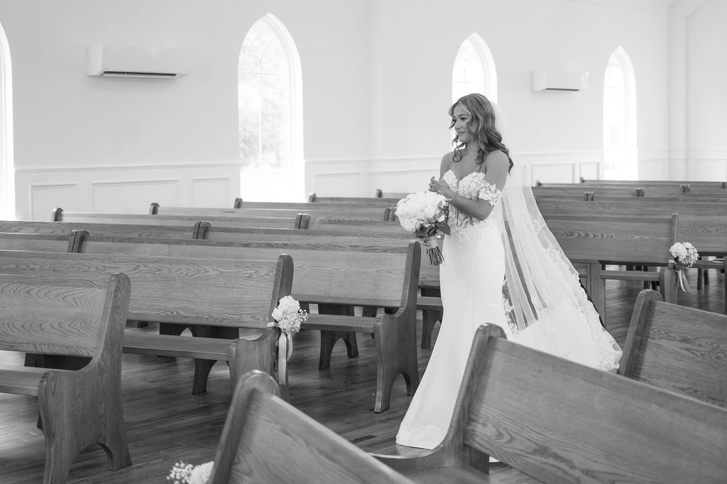 Bride in white wedding gown walking down church aisle with wooden pews in black and white photo.