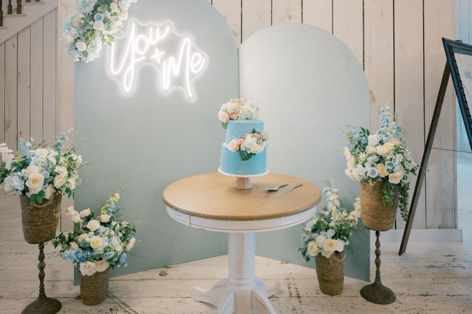 Blue wedding cake with white florals on round table with neon you and me sign backdrop.