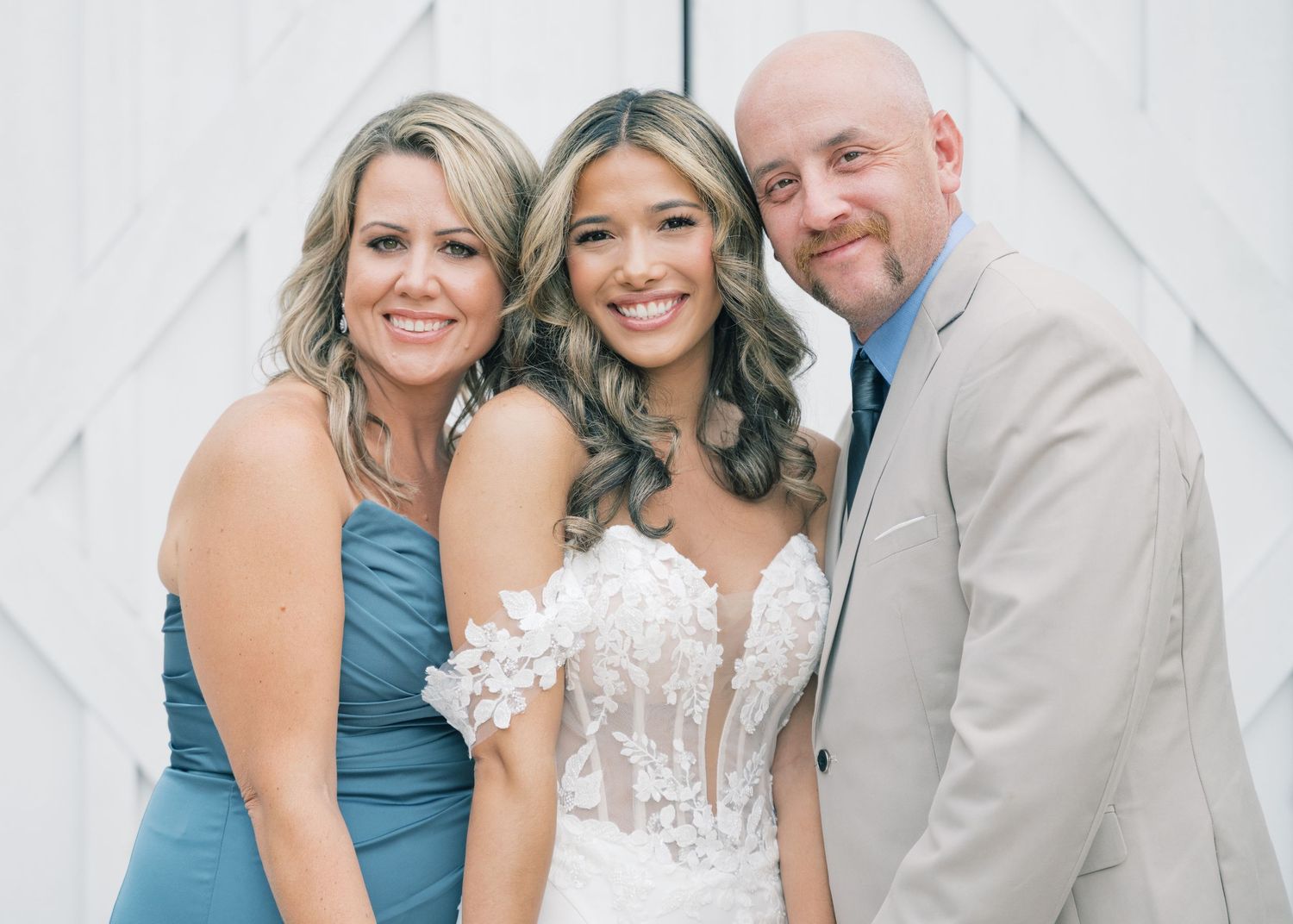 Bride in strapless lace gown posing with two smiling guests in blue and tan attire at wedding venue.