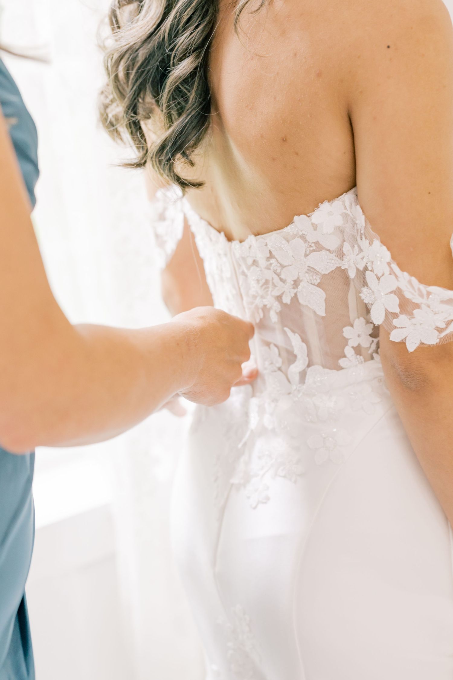 Close-up of bride's lace floral applique wedding dress being fastened by a helper before the ceremony.