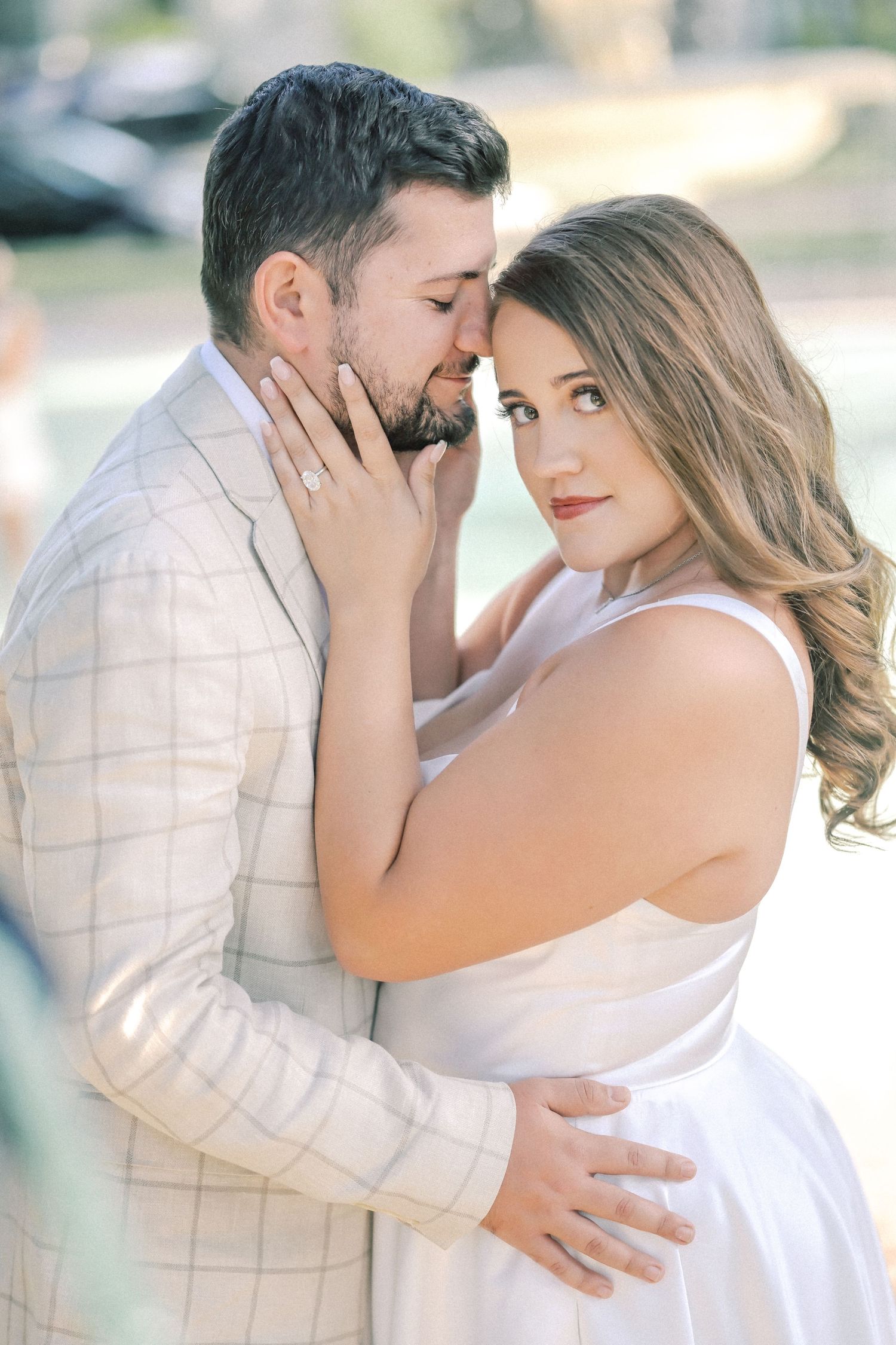 Woman in a white dress gazes at the camera while cradling her fiancé's face, her engagement ring visible in a soft outdoor portrait.