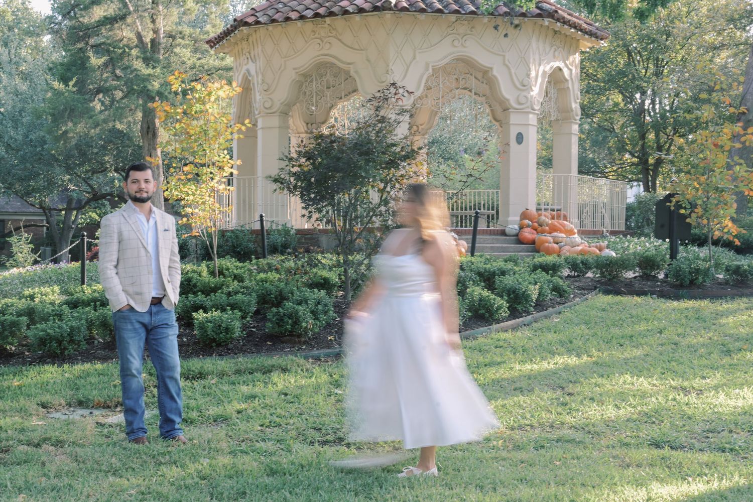 Man in a blazer and jeans stands on a lawn as a woman in a white dress twirls in motion blur near a garden gazebo.