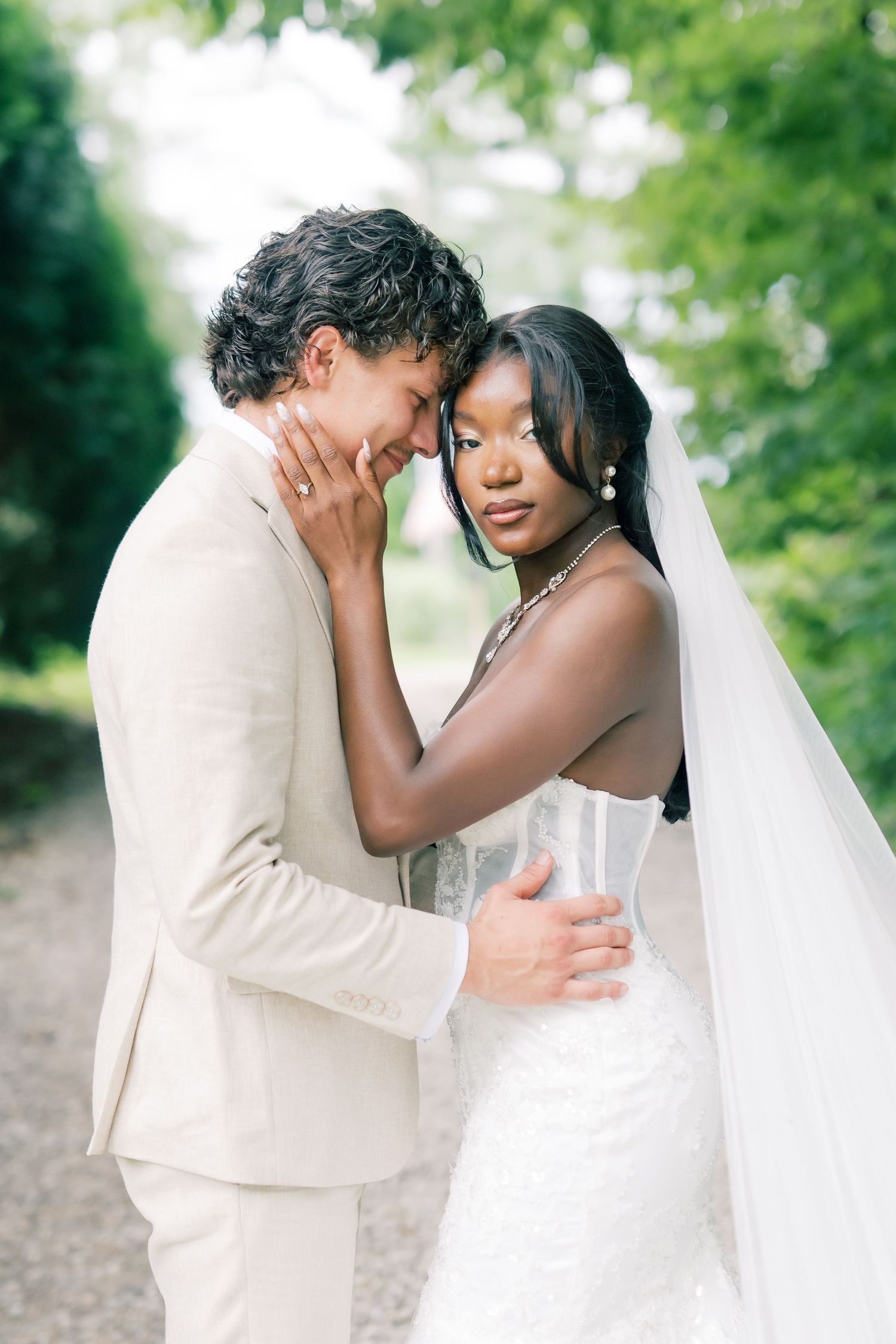 Interracial couple sharing a romantic moment on their wedding day, bride in white gown with veil, groom in beige suit.