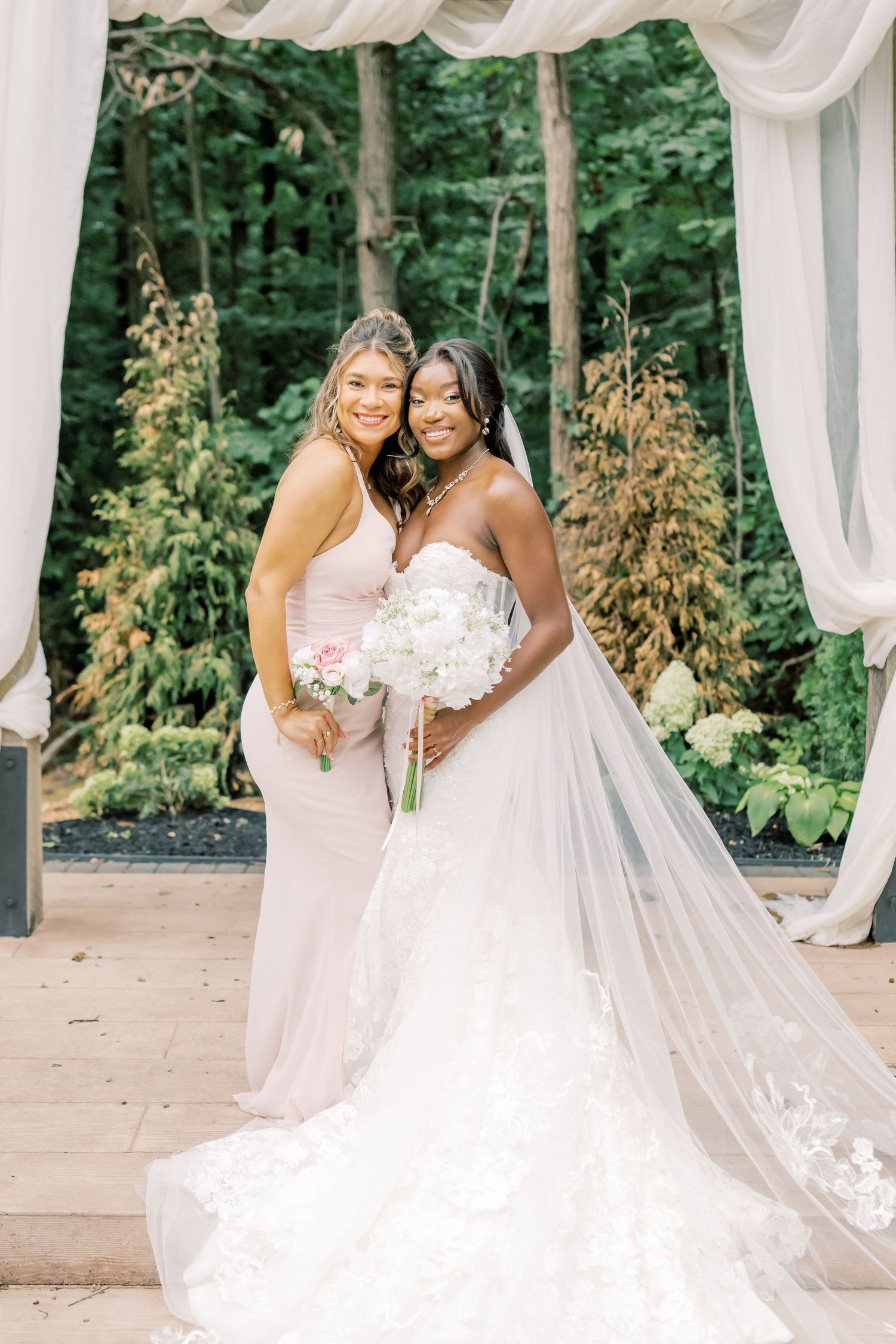 Bride in strapless lace gown holds white bouquet beside bridesmaid in blush dress at outdoor altar.