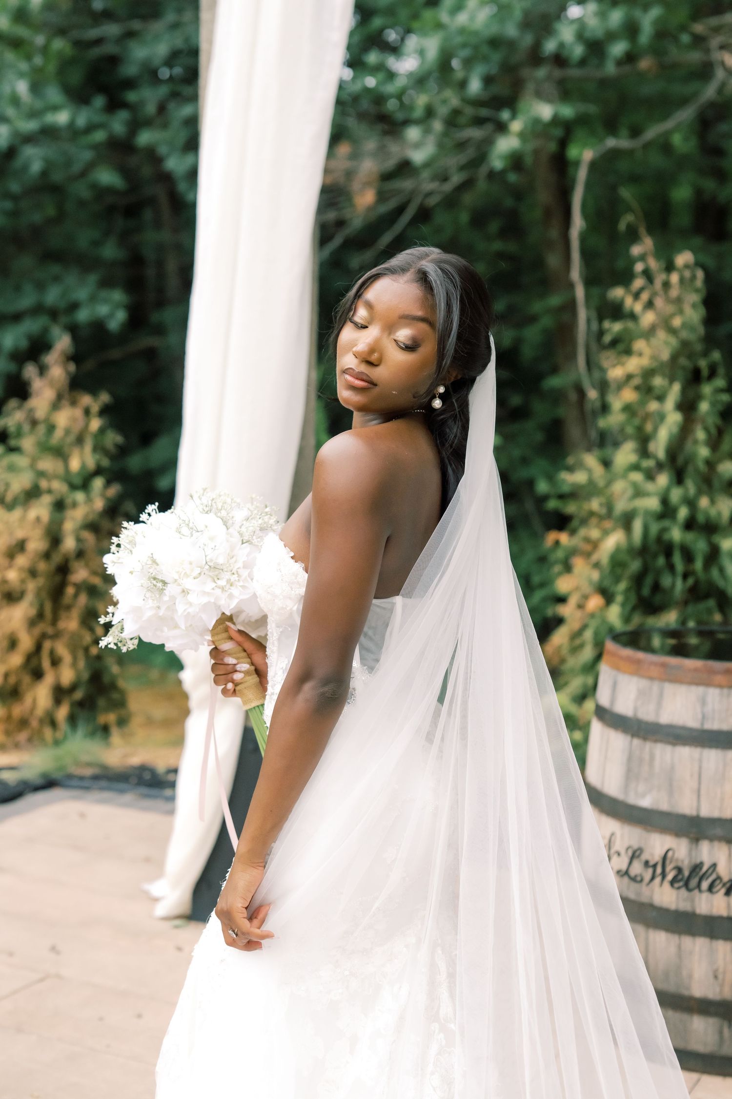 Elegant Black bride in white lace gown and veil holds white bouquet outdoors at a garden wedding venue.