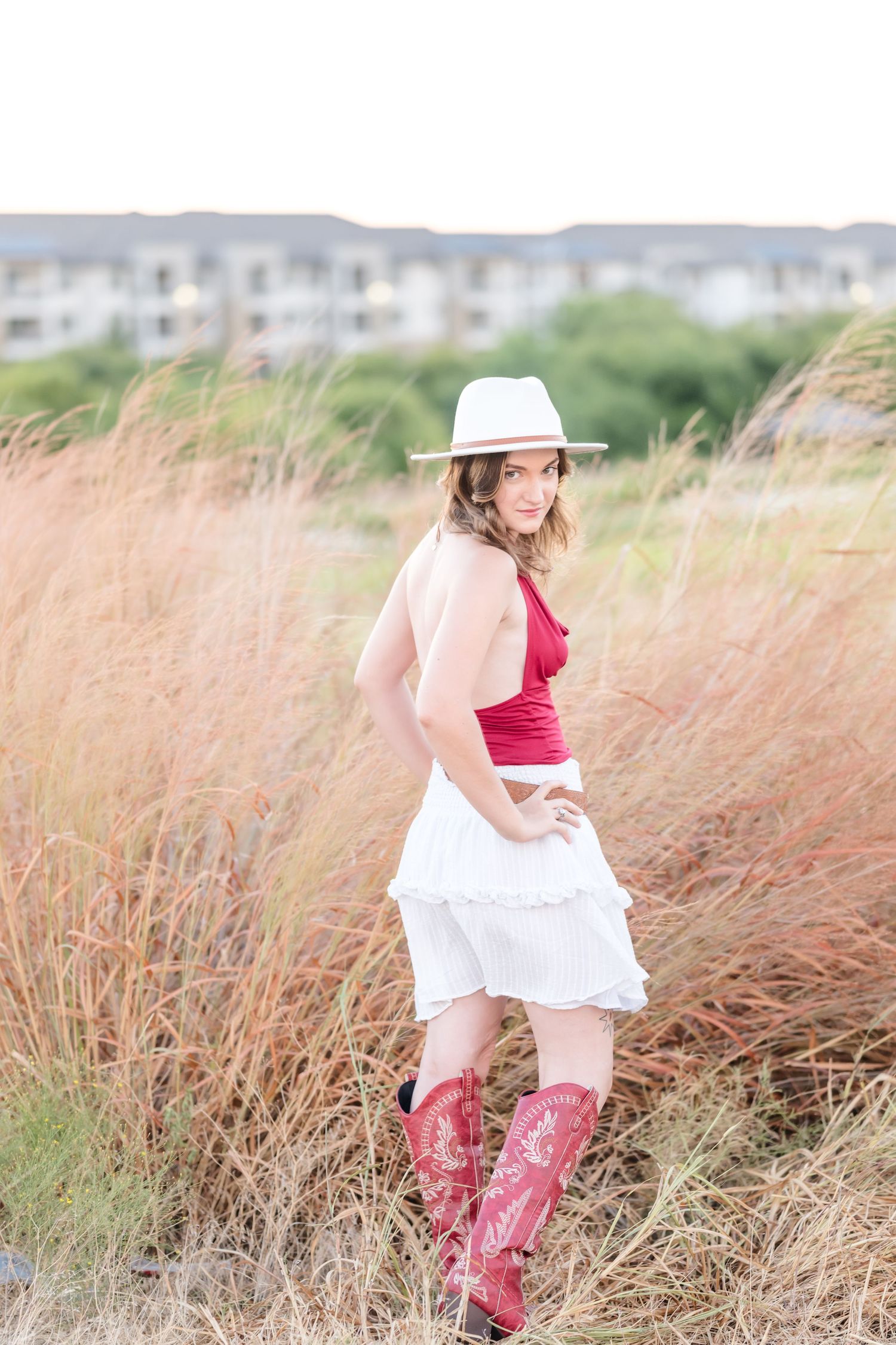 Young woman in white hat, red top, white ruffled skirt, and red cowboy boots posing in tall grass field.