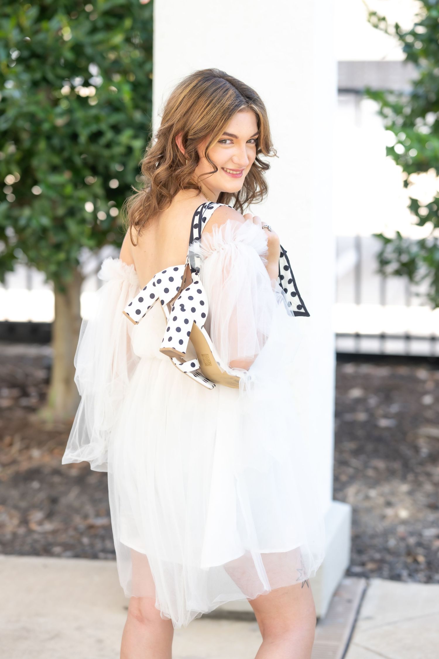 Woman in white tulle dress with polka dot bow, posing outdoors near greenery.