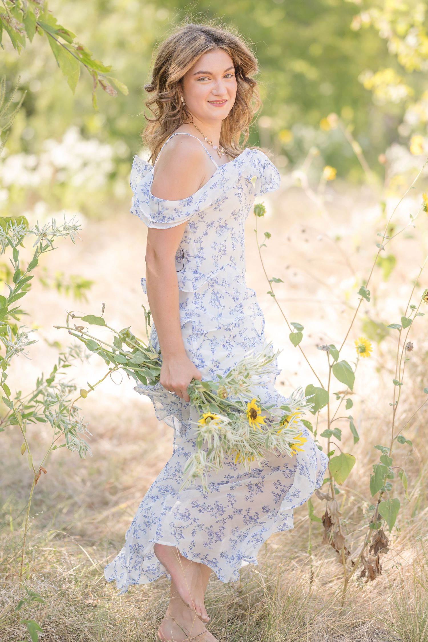 Young woman in floral off-shoulder dress holding sunflowers in a sunlit meadow.