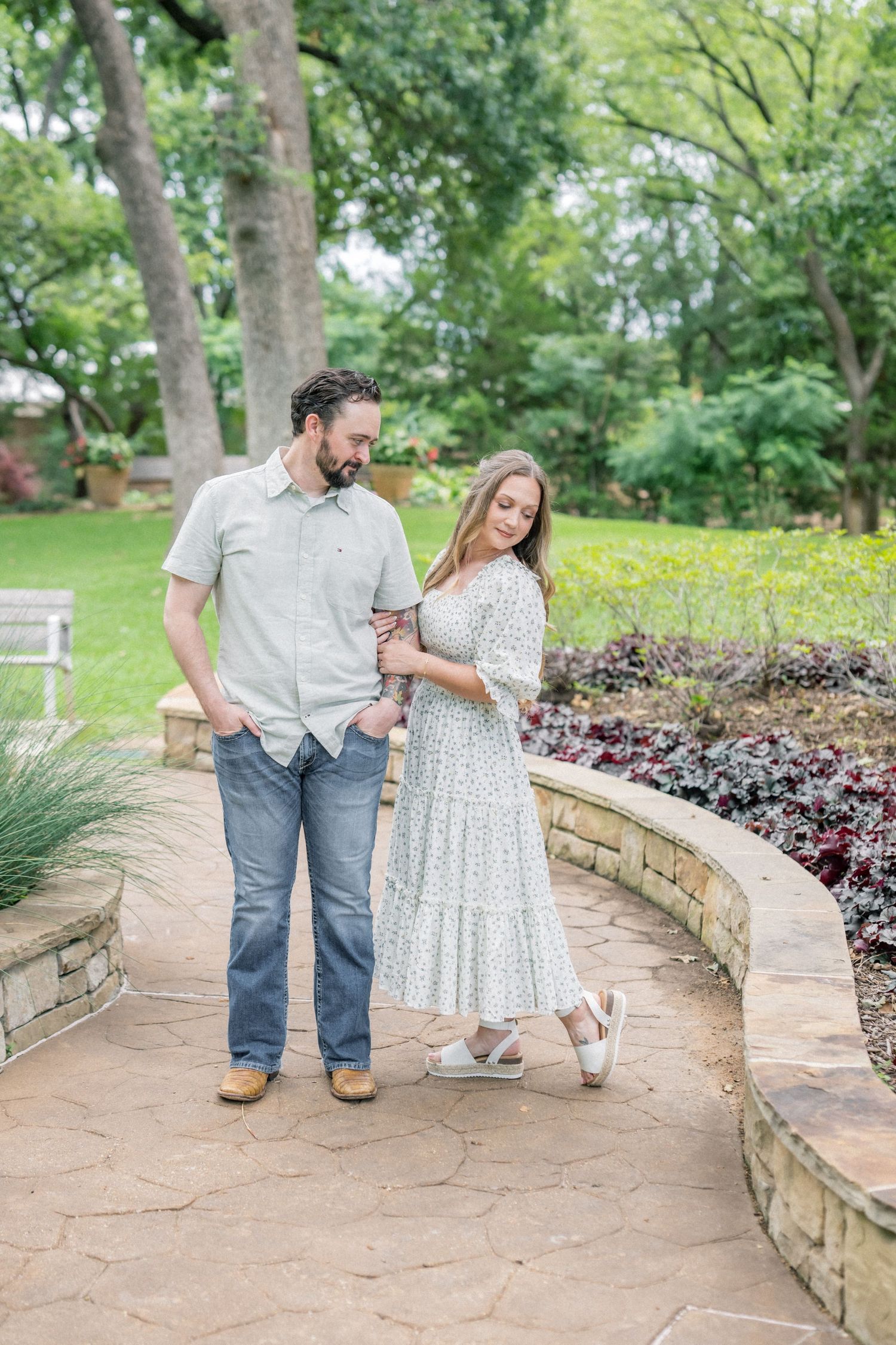 Couple walking hand-in-hand along a stone garden path surrounded by lush greenery and flowers.