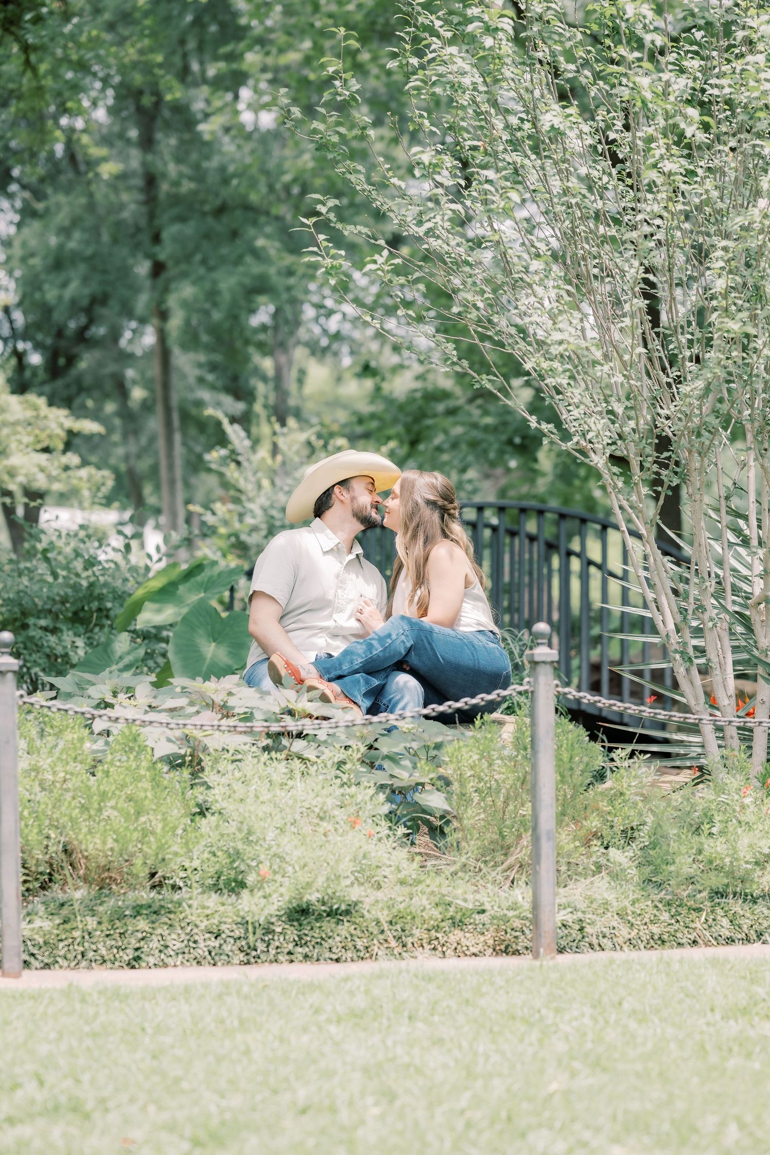 Couple sharing a kiss while sitting on a bench surrounded by greenery, man wearing a straw cowboy hat.