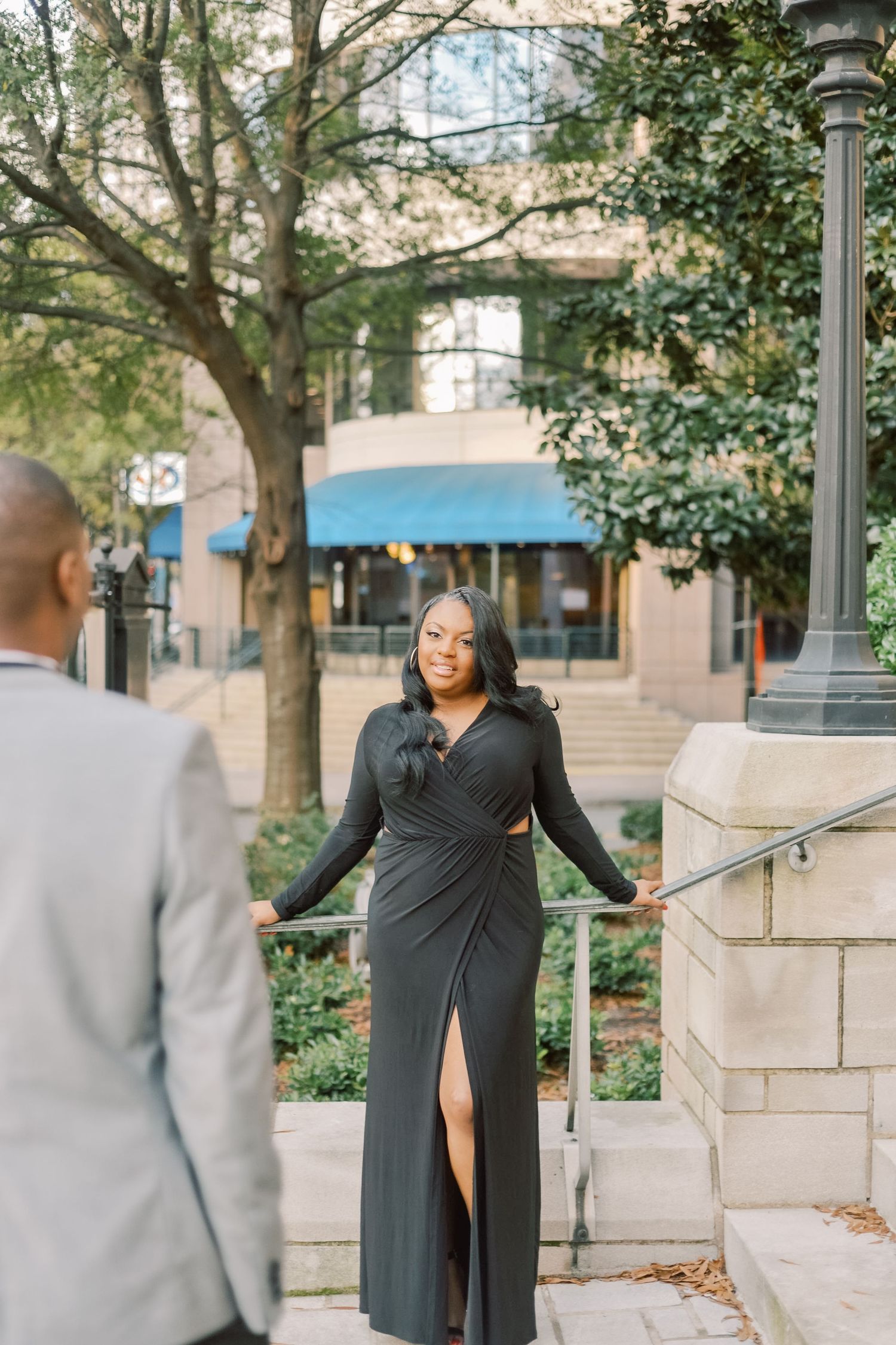 Woman in black wrap dress poses on urban staircase railing while man in gray suit approaches.