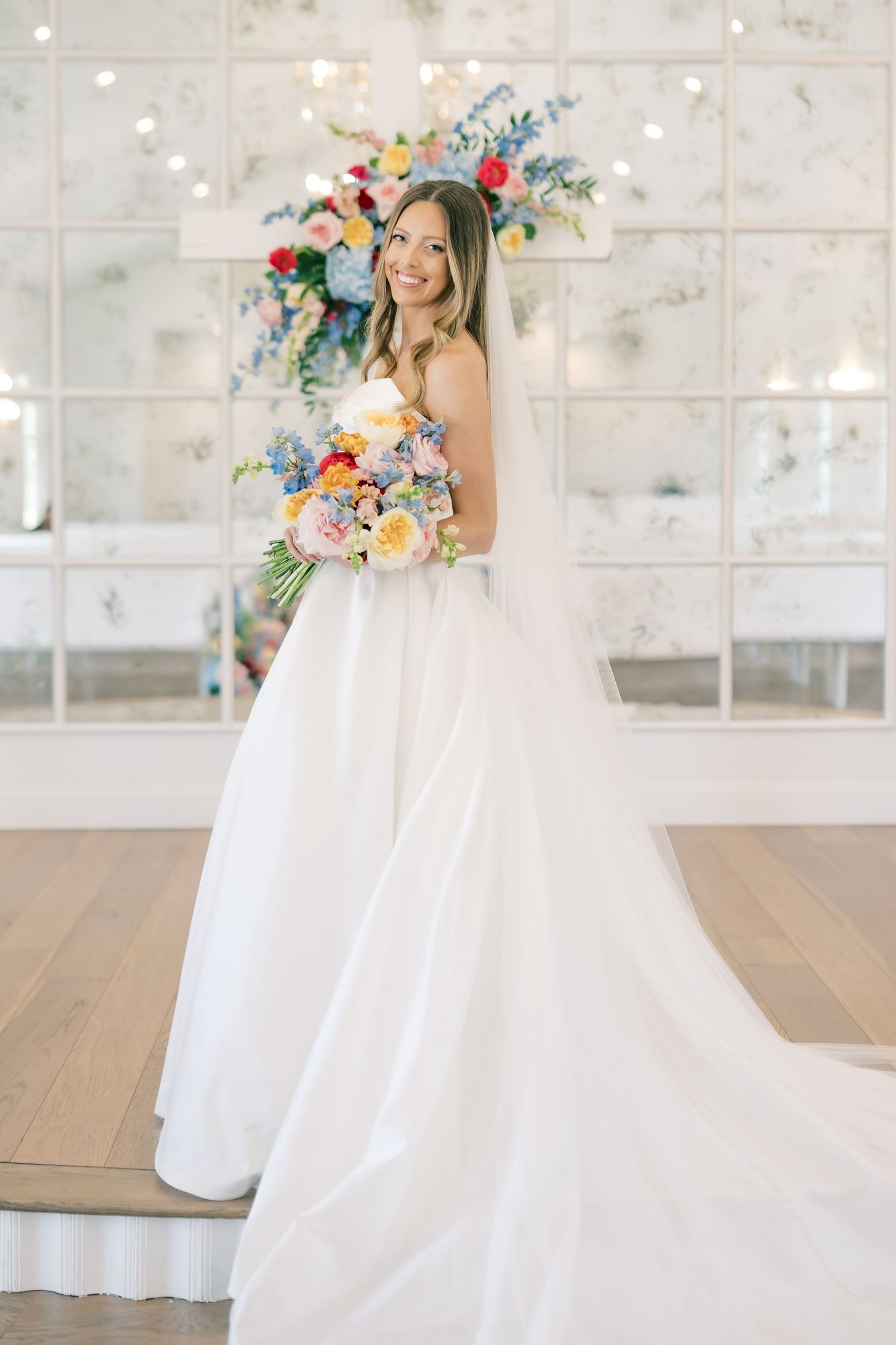Bride in strapless white ballgown holding colorful bouquet, smiling in elegant mirrored venue.