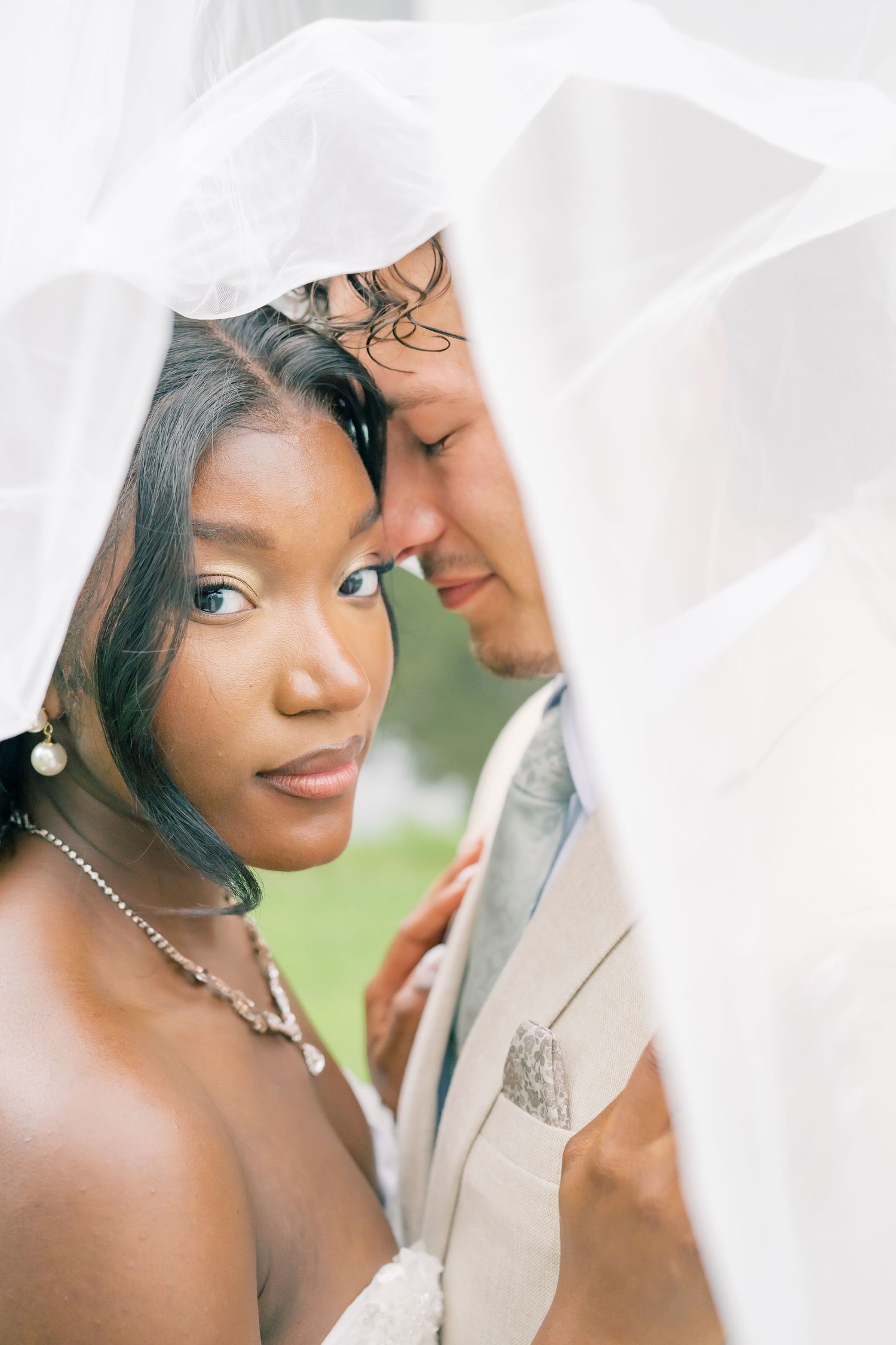 Bride and groom share an intimate moment under a white veil, touching foreheads in a romantic outdoor wedding portrait.