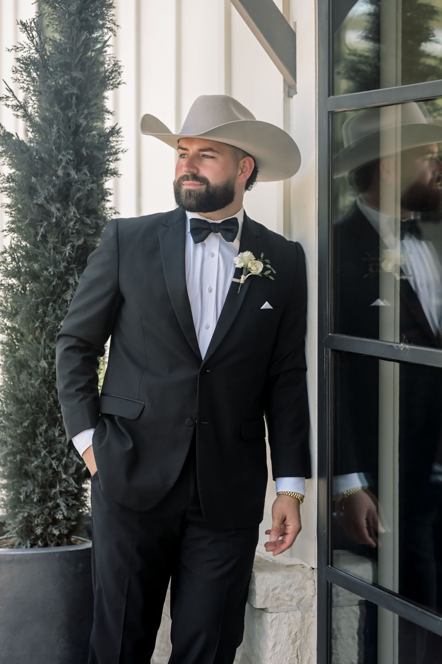 Dapper groom in black tuxedo and white cowboy hat stands confidently outside wedding venue.