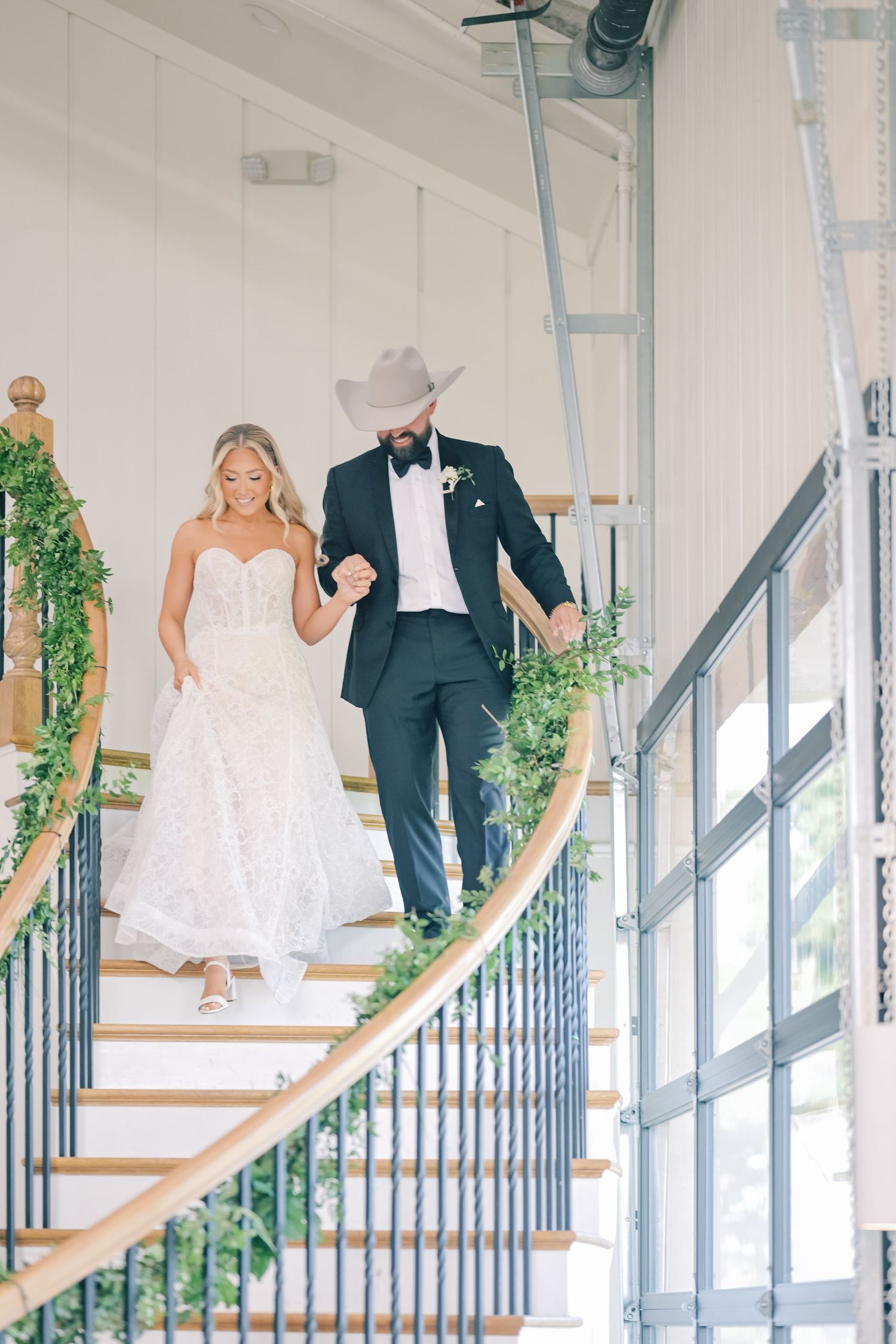 Smiling bride and groom in cowboy hat descend spiral staircase decorated with greenery garland.