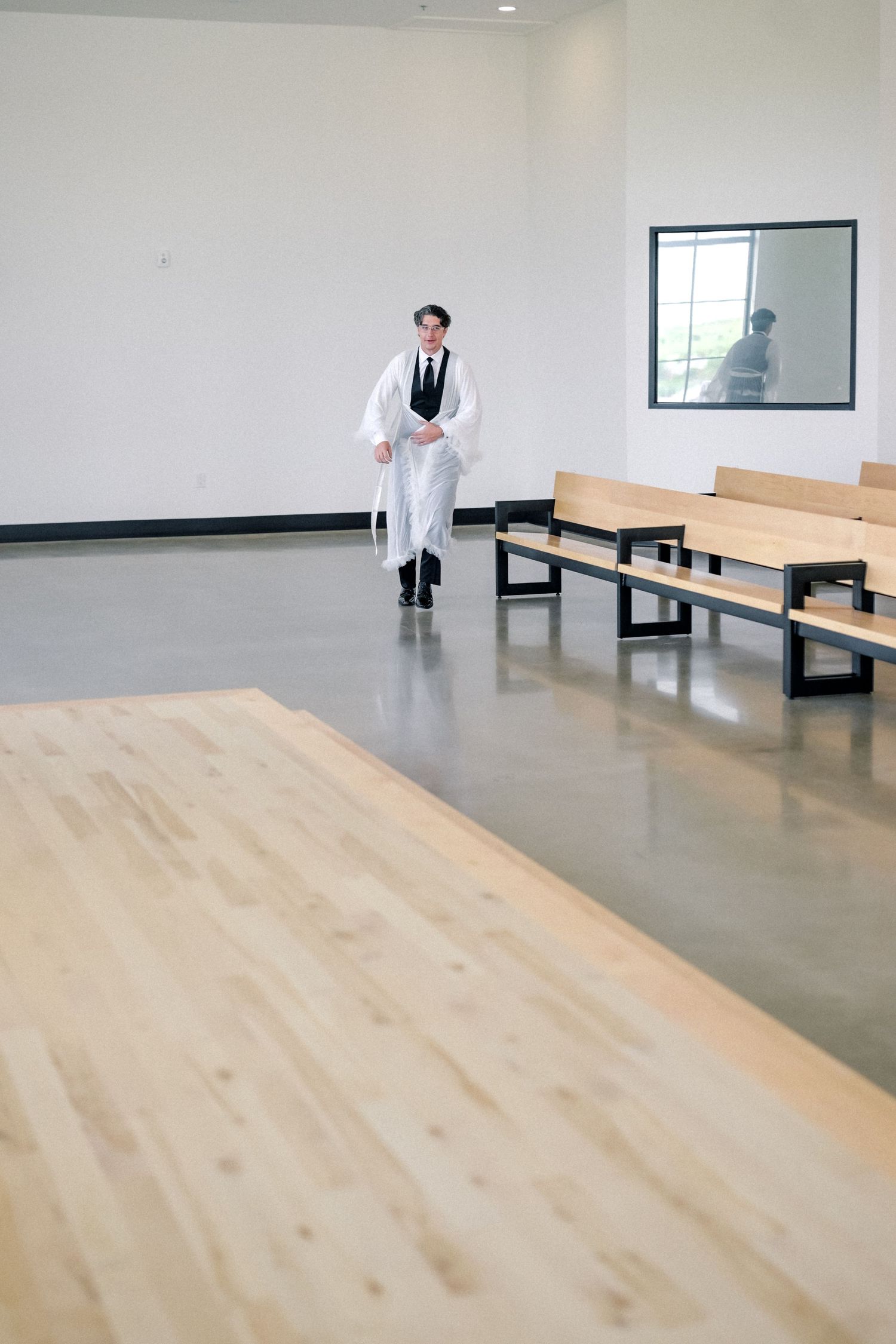 A rabbi in white robes walks between wooden pews in a modern minimalist wedding chapel with polished concrete floors.