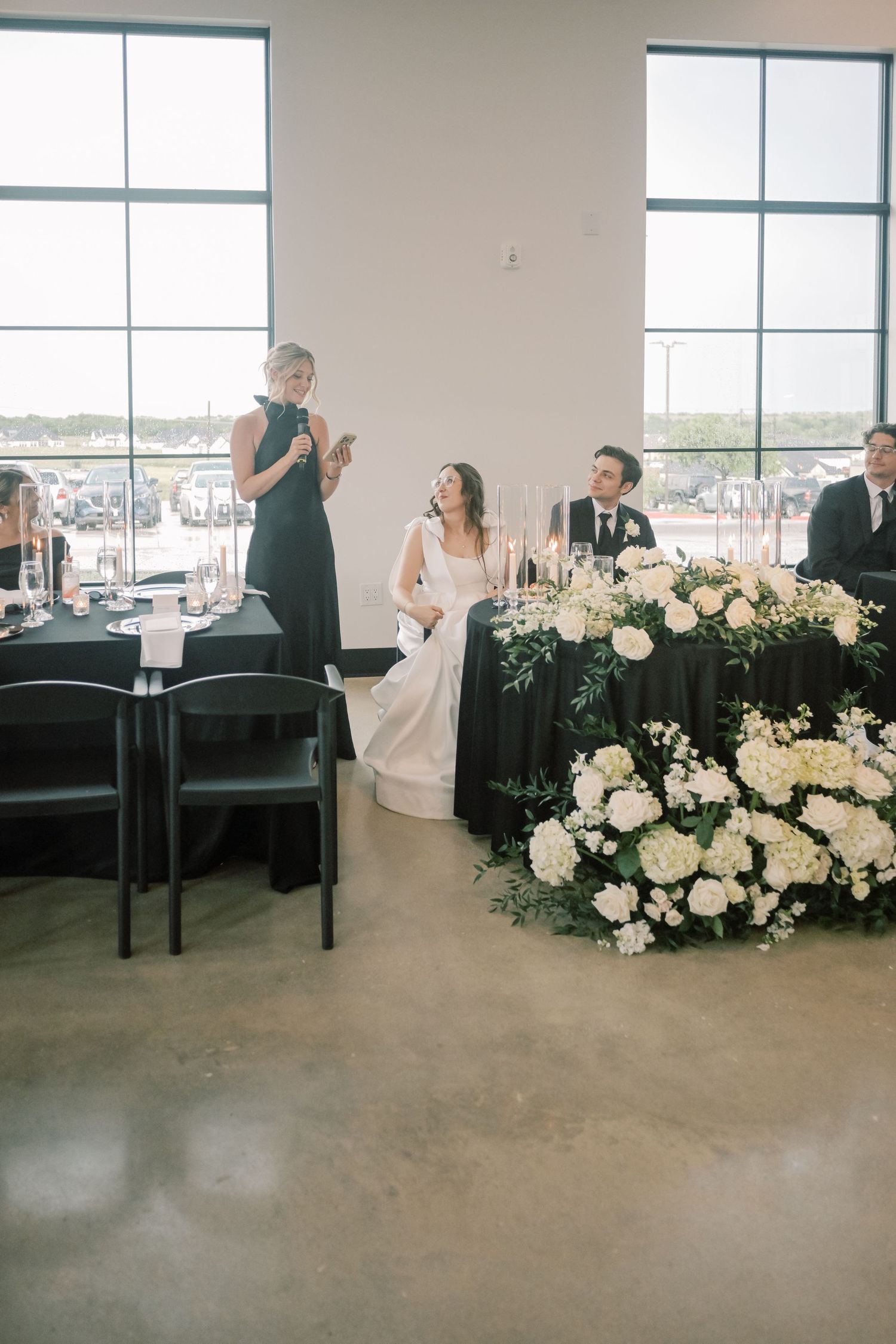 Bridesmaid giving a toast speech while bride and groom smile at a floral-decorated sweetheart table.