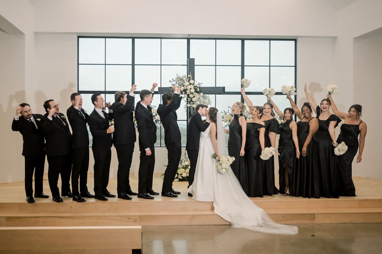 Wedding party in black attire celebrates with the bride and groom at the altar in a modern church.