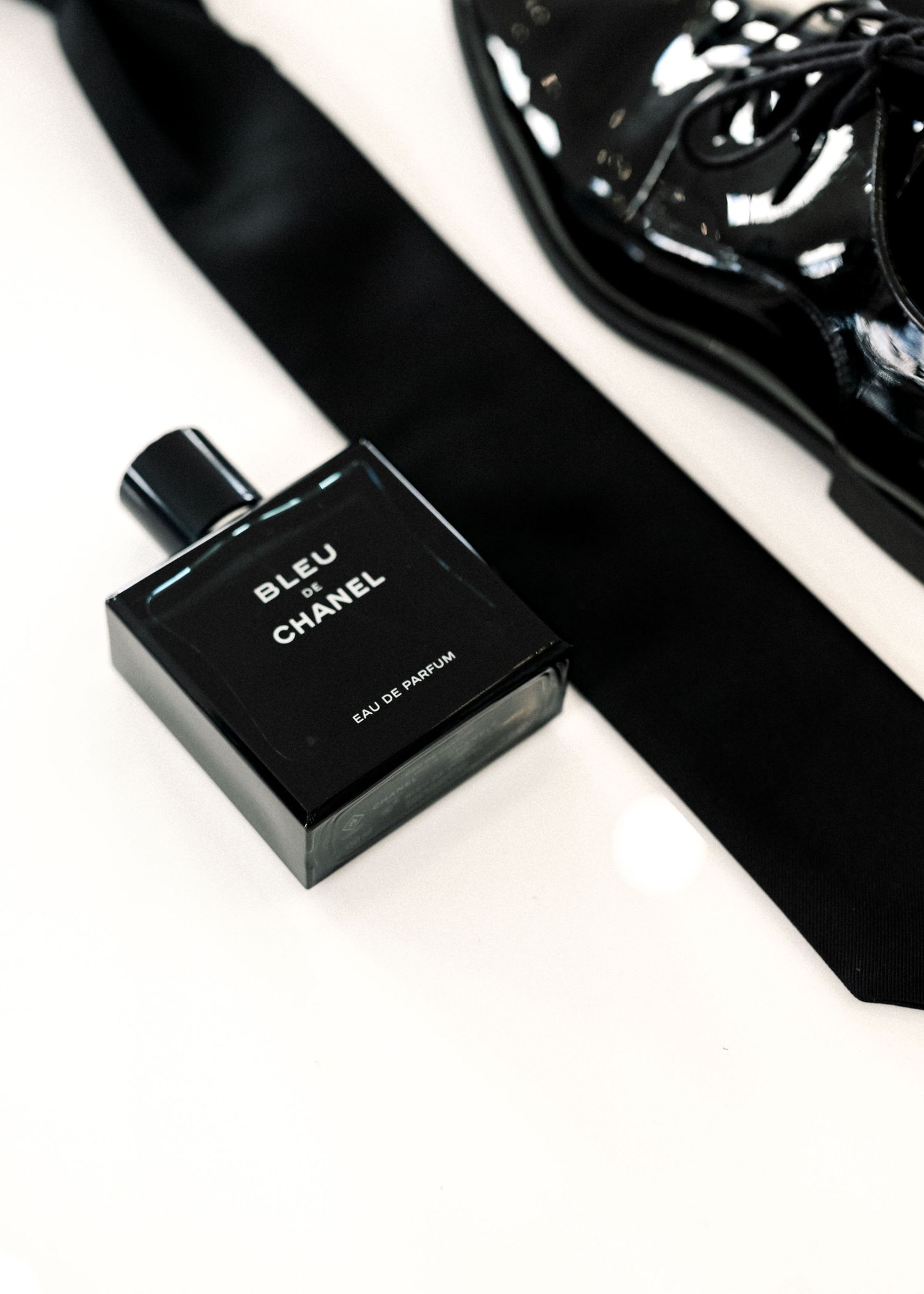 Bleu de Chanel Eau de Parfum bottle beside a black silk tie and patent leather shoe on white surface.