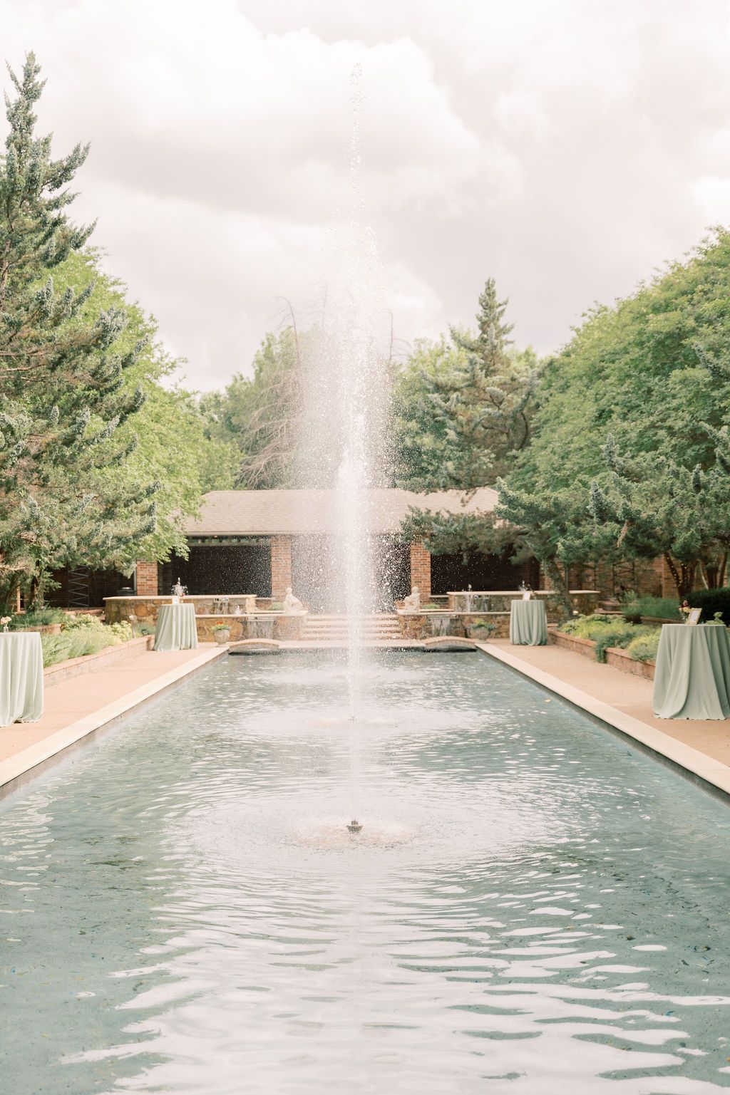 Elegant outdoor fountain centerpiece in a long reflecting pool surrounded by lush green trees and draped event tables.