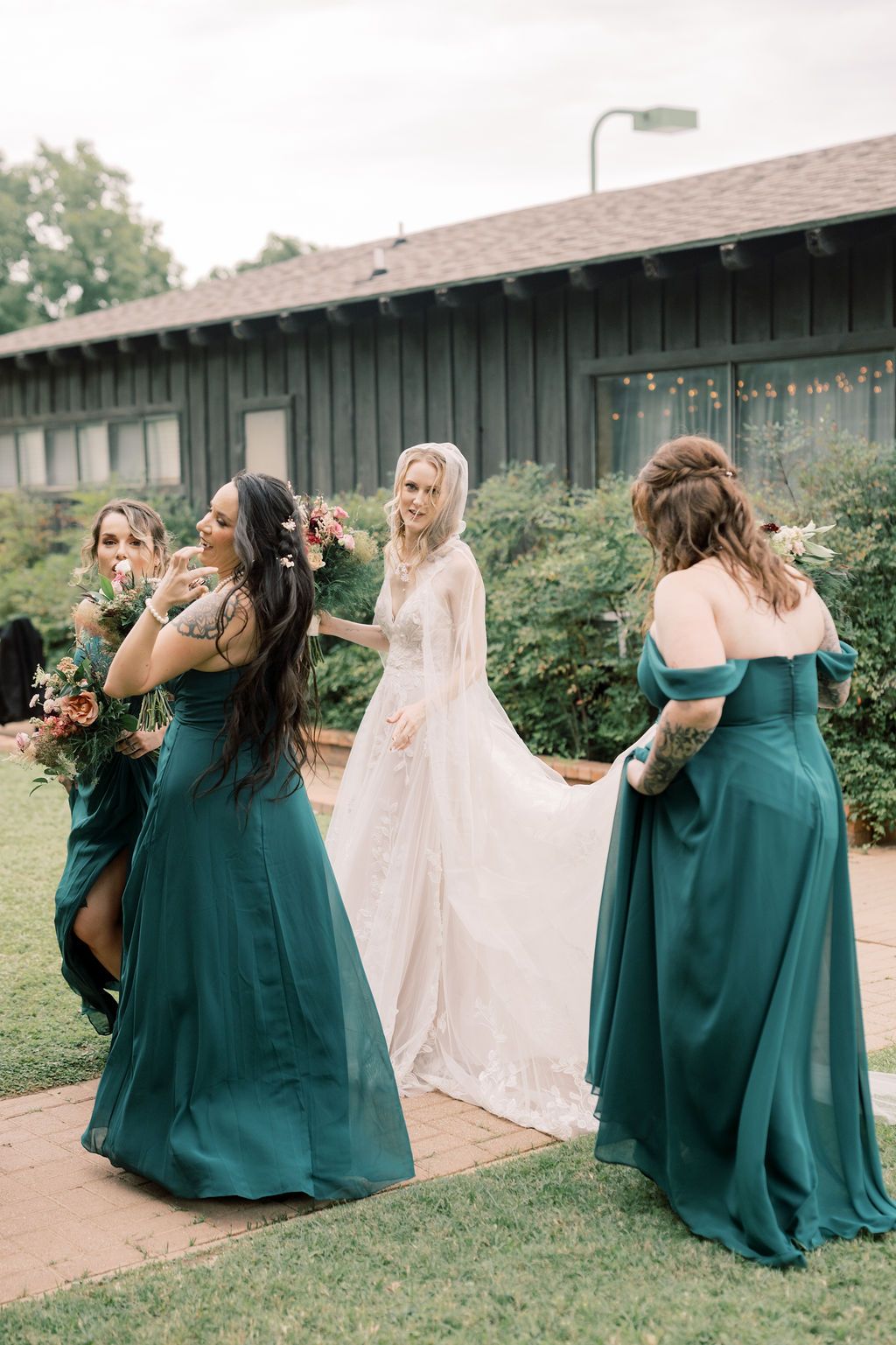 Bride in white lace gown with bridesmaids in emerald green dresses outside rustic venue.