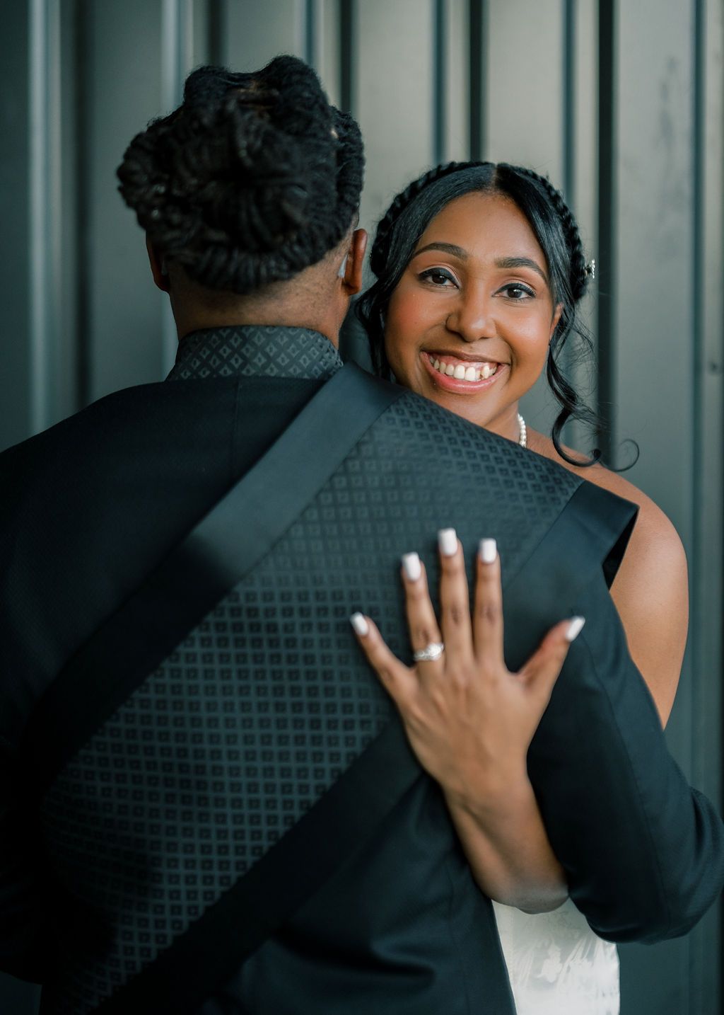 Smiling bride in white dress hugs groom in black suit, showing off her engagement ring during a wedding photo shoot.