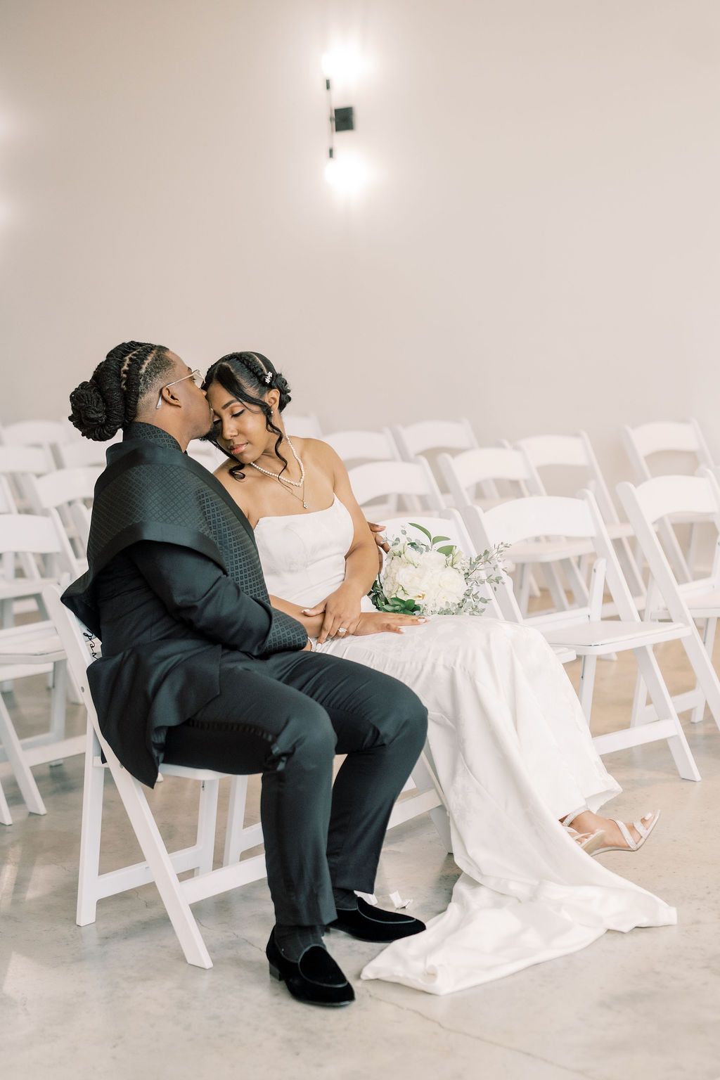 A bride and groom sit intimately together on white chairs in an empty wedding ceremony venue, sharing a tender moment.