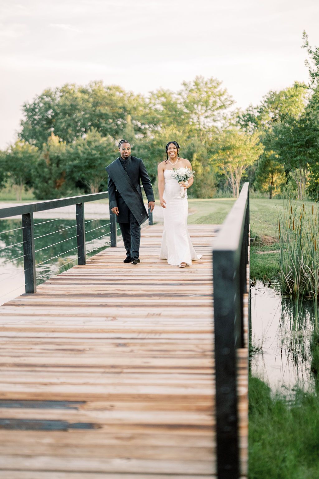 Bride and groom walk hand-in-hand across wooden bridge at outdoor wedding venue.