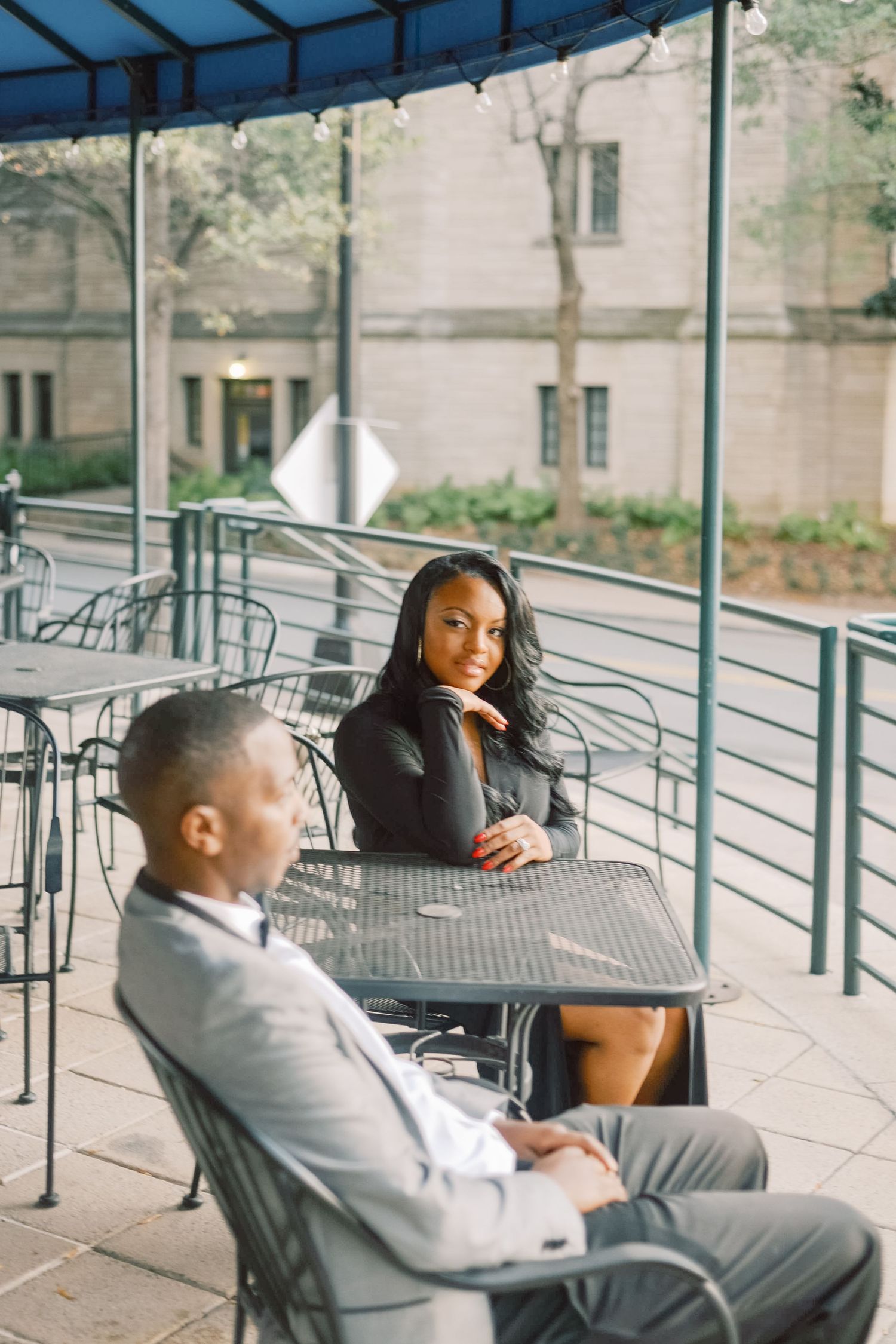 Stylish Black couple at outdoor cafe patio, woman in black dress smiling at man in gray suit seated nearby.