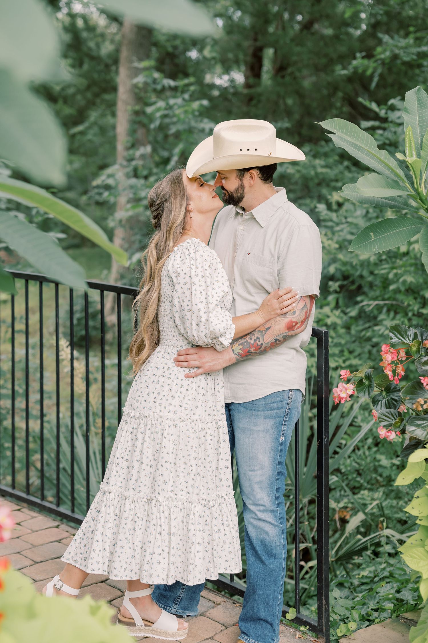 A couple shares a kiss on a garden balcony, man in cowboy hat and jeans, woman in white floral dress.