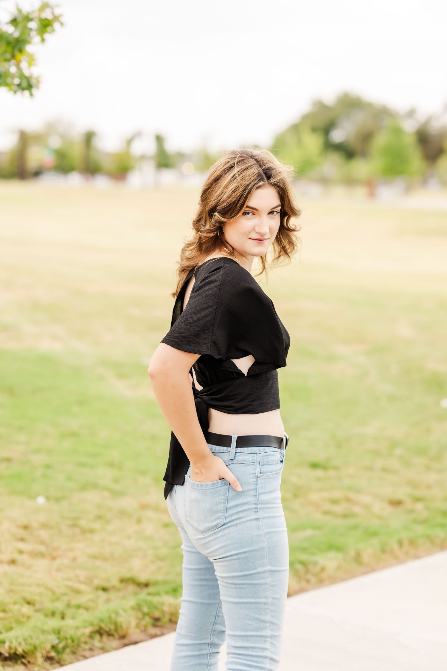 A young woman in a black top and light blue jeans poses outdoors in a park, glancing back over her shoulder.