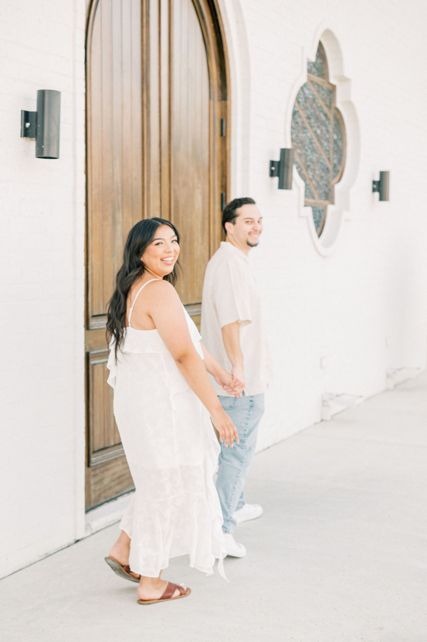 A couple laughs together while posing in a bright white corridor with rustic wooden doors.