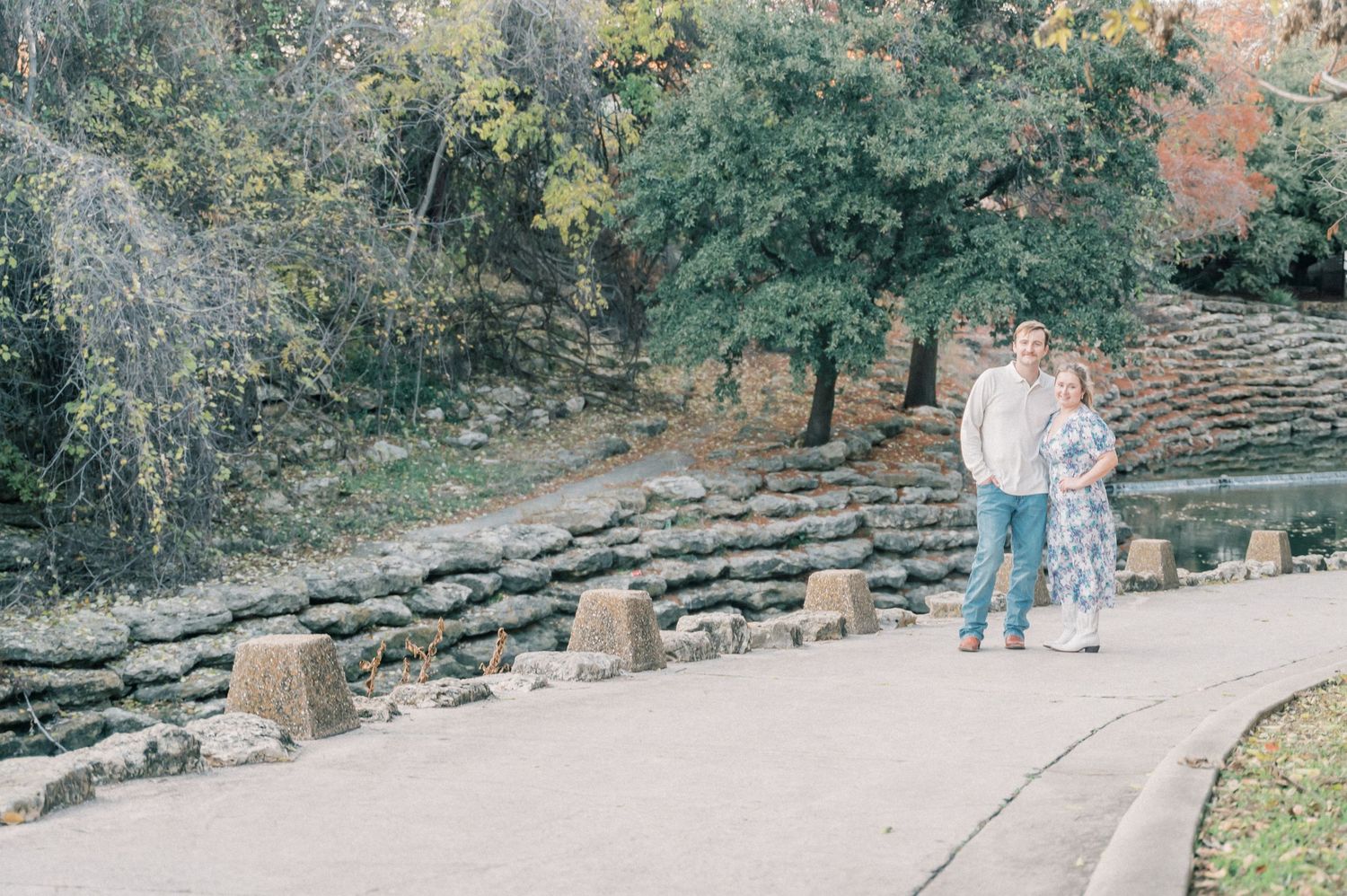 A couple stands together on a stone path next to rustic amphitheater seating in a wooded outdoor setting.