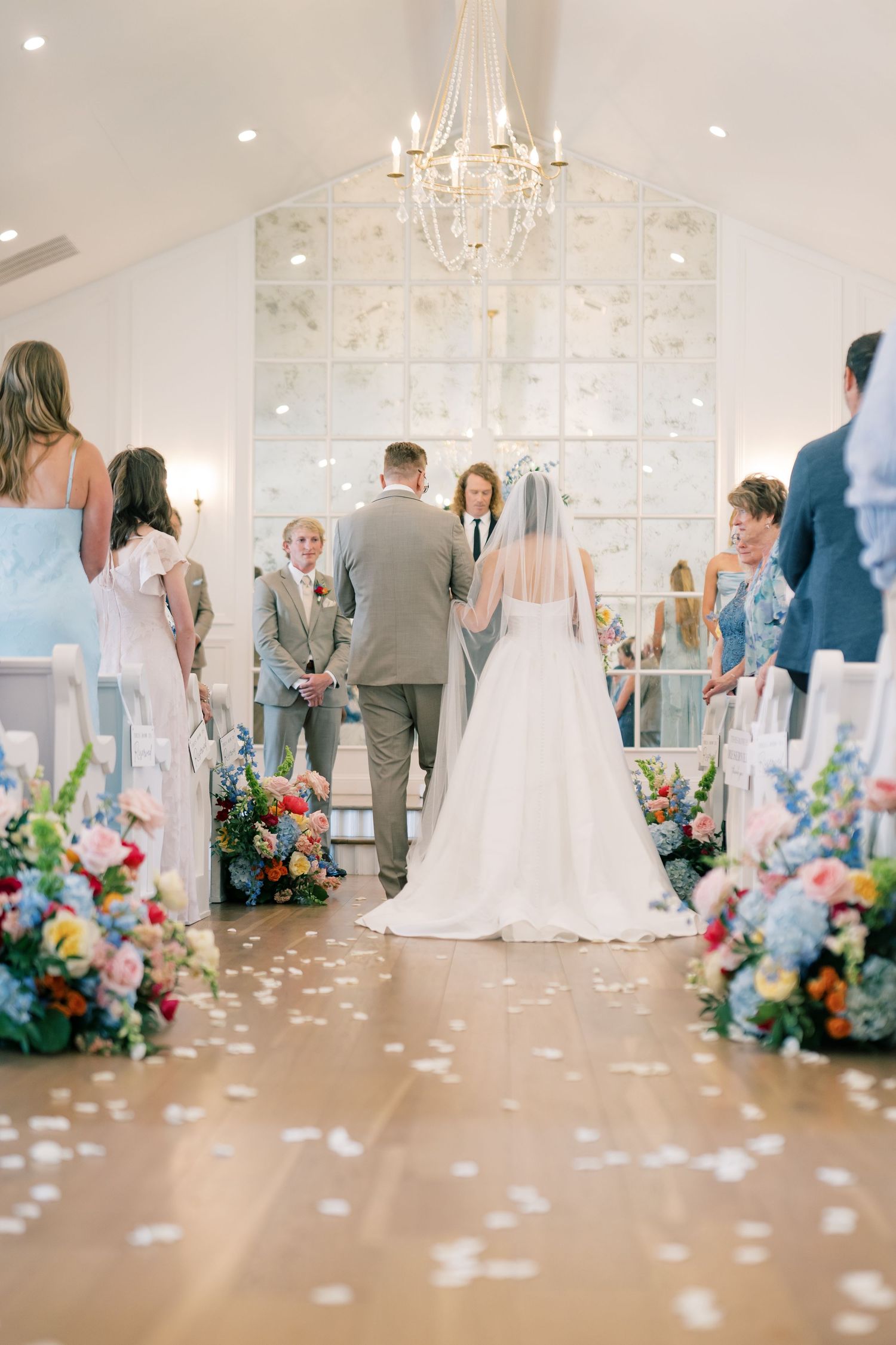 Bride and groom at altar in bright chapel with colorful floral aisle arrangements and chandelier.