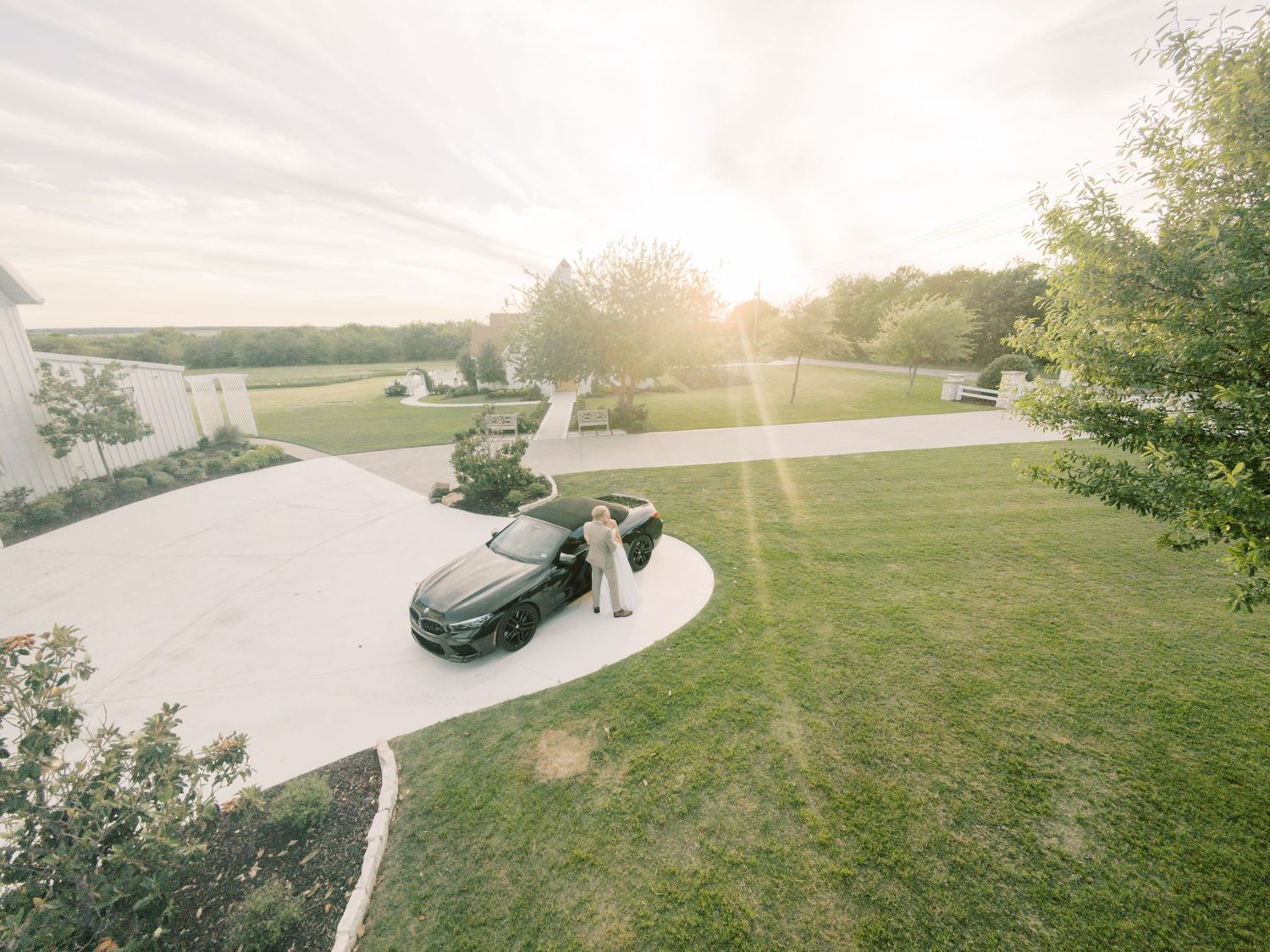 Aerial view of a person standing by a dark luxury car in a circular driveway surrounded by lush green lawn at sunset.